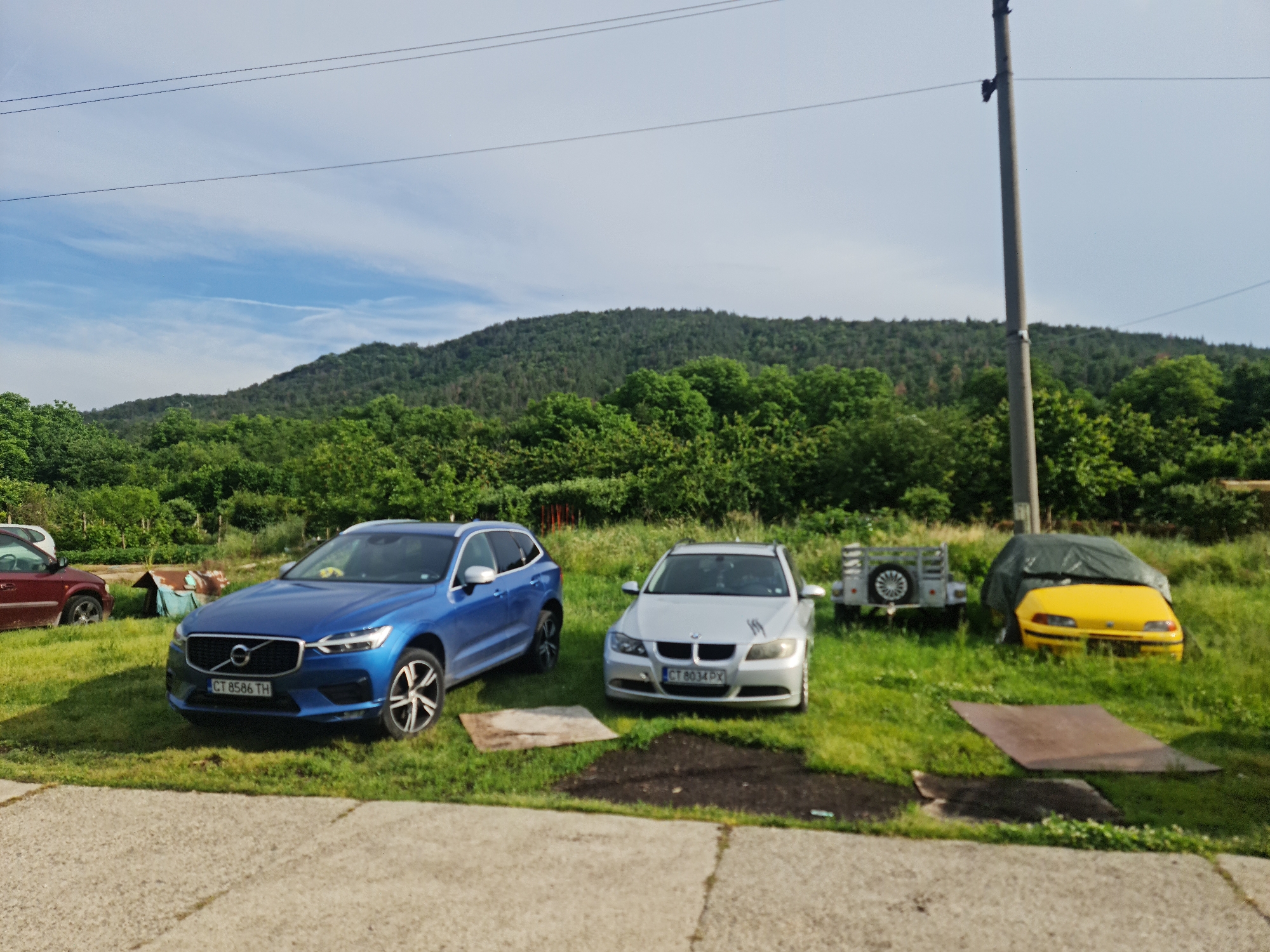 This outdoor scene in Buzovgrad, Bulgaria, depicts a parking or storage area for several vehicles, set against a backdrop of lush green hills and a partly cloudy blue sky. The time of day appears to be bright daylight.
In the foreground, a concrete surface gives way to a grassy expanse. A prominent blue Volvo SUV, with license plate "CT 8586 TH", is parked on the grass on the left side of the frame, facing slightly towards the viewer. To its right, a silver BMW sedan (likely a 3-series touring model) is also parked on the grass, displaying the license plate "CT 8034 PX". Further right, a small, light-colored utility trailer with a spare tire attached is visible. Next to the trailer, a yellow car, possibly a Fiat Punto, is largely covered by a dark tarp, with only its front fascia and headlights exposed. A dark red car is partially visible on the far left edge of the image.
The background is dominated by a tree-covered mountain range, indicative of a rural or semi-rural setting. Overhead, thin power lines stretch across the sky, connected to a tall utility pole on the right side of the frame. The ground is primarily green grass, with a few patches of bare soil or disturbed earth, and two flat, rectangular objects (possibly metal sheets) are placed on the grass in the mid-ground. No people are present in the image. The weather is clear and sunny, suggesting pleasant conditions.