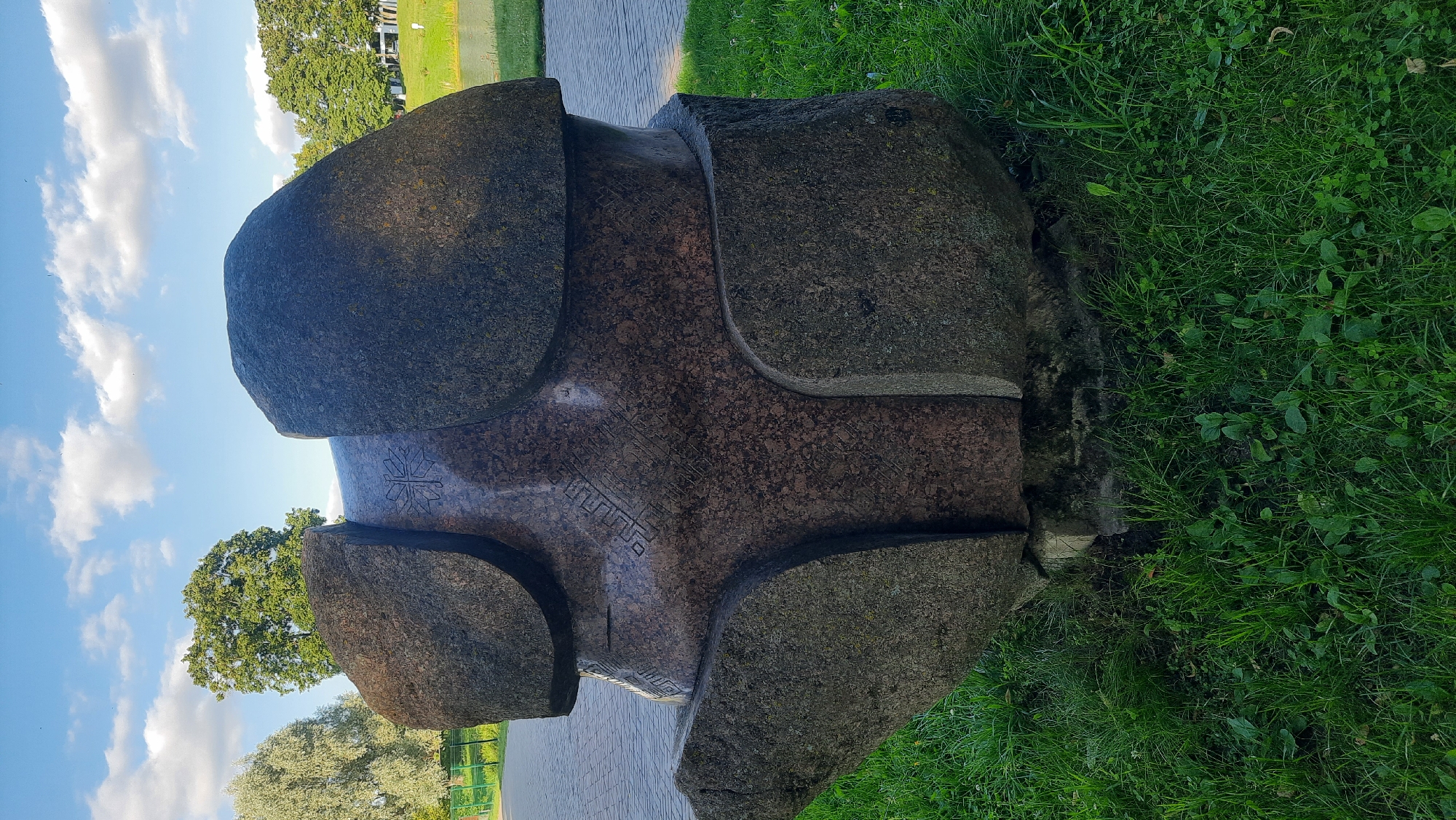 The image depicts a large, abstract stone sculpture situated outdoors on a grassy area. The sculpture is composed of several rough-hewn stone pieces, some of which appear to be granite, fitted together to form a rounded, somewhat organic shape. There are carved geometric patterns on the central, smoother part of the sculpture.
Behind the sculpture, a paved pathway is visible, suggesting a park or public space. Further in the background, there are trees and a body of water, possibly a pond or lake. The sky is blue with scattered white clouds.
This sculpture is located in the city of Smarhon, Belarus. It is situated near the Smarhon City Culture Palace. The area surrounding the sculpture is well-maintained with green grass and a paved walkway. The overall impression is of a public art installation in a recreational area.