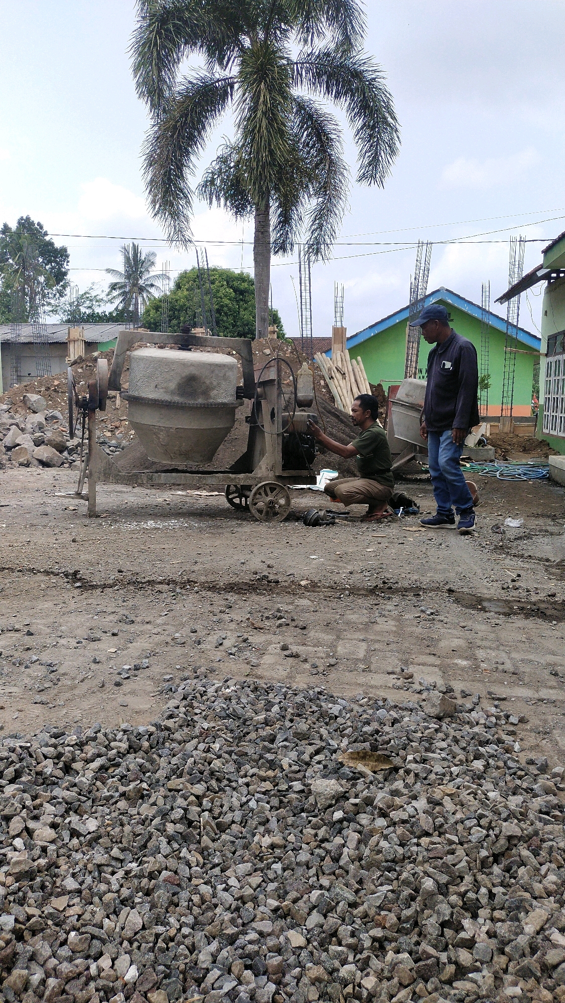 In Klungkung, Indonesia, a construction site is depicted. A concrete mixer, a staple on such sites, is centrally positioned. Two men are engaged with it. One, wearing a dark cap and blue jeans, stands to the right, observing. The other, in a khaki shirt and brown pants, crouches beside the mixer, appearing to be adjusting or repairing it. The ground is covered in gravel and dirt, indicative of ongoing construction. In the background, unfinished buildings and construction materials like lumber and rebar suggest the development of new structures. A tall palm tree stands prominently, its fronds reaching towards the overcast sky, adding a tropical element to the otherwise industrial scene.