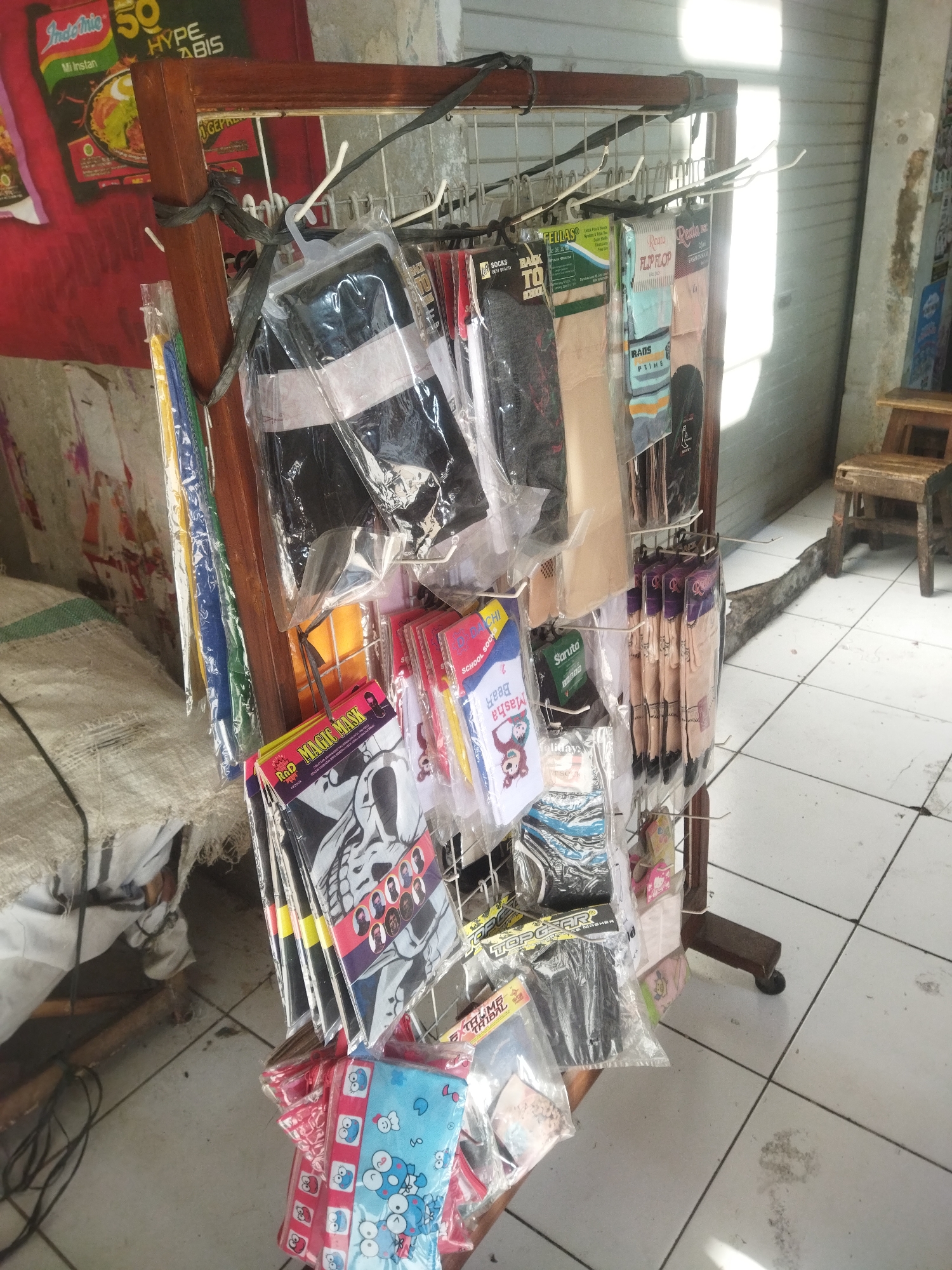 The image shows a street-side stall in Sukoharjo, Indonesia, displaying various items of clothing and accessories, primarily socks and masks. The stall is constructed with a wooden frame and a metal rack, from which numerous items are hung or displayed on shelves. The items are mostly packaged in plastic.

On the left side of the stall, there are bundles of colored fabric strips, possibly for sale. Further to the right, multiple pairs of socks are displayed, some in plastic packaging with branding such as "FELLAS," "Roms," and "Saruta." There are also several face masks, including some with skull graphics and cartoon characters.

The background suggests an outdoor setting, with a tiled floor and a large, grey, corrugated metal door or wall behind the stall. Sunlight streams in from the right, casting strong shadows. To the far right, a rustic wooden stool is visible. The general impression is that of a small, informal retail operation catering to passersby.