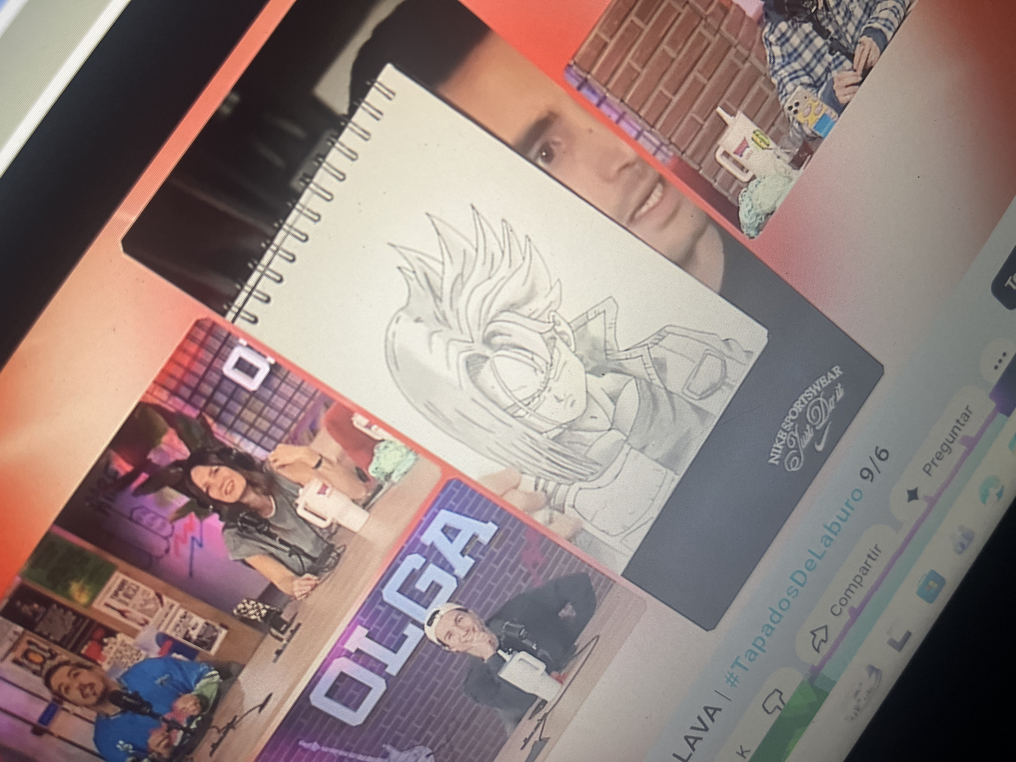 The image shows a screen displaying a video feed or stream. In the foreground, a sketchbook is open, featuring a pencil sketch of a character with spiky hair, likely from an anime or manga. The sketchbook rests on a table. Behind the sketchbook, on the screen, are images of people. To the left, there are two individuals sitting at a desk, seemingly participating in a broadcast or podcast, with microphones in front of them. One person wears a blue jacket, and the other a grey top. The word "OLGA" is visible in large letters on a background behind them. Further to the right on the screen, a man's face is visible, looking towards the camera, and behind him, a brick-like pattern is present. Another person in a plaid shirt is also partially visible on the right side of the screen. The overall scene suggests a digital media setup, possibly a studio or home recording environment. There is no discernible information within the image that indicates the location is the city of Santa Ana, Venezuela.
