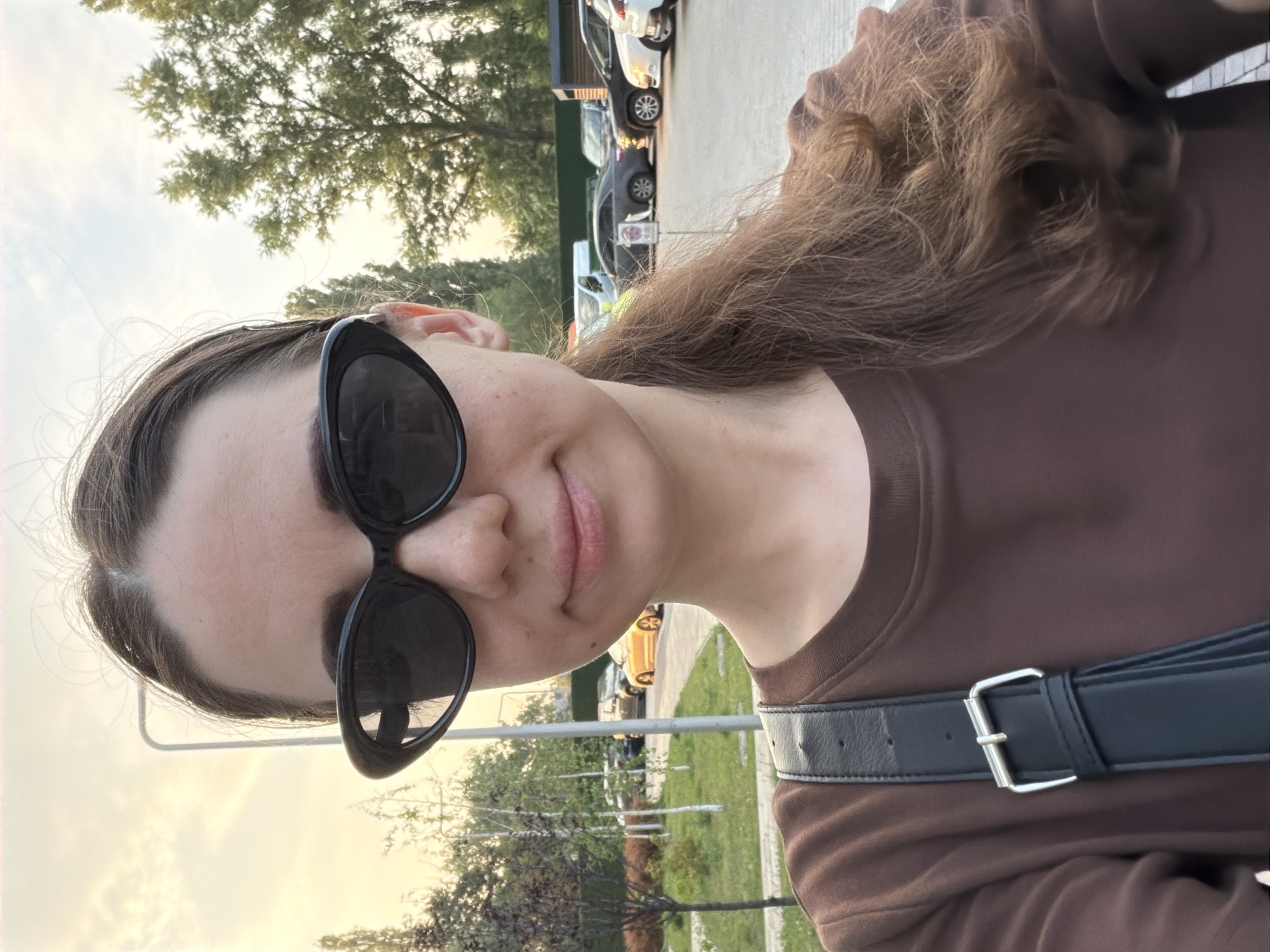 A woman with brown hair, wearing a brown sweatshirt and black sunglasses, stands outside with a blurred background of trees and parked cars. The scene appears to be a street or parking lot, possibly in Kyiv, Ukraine, given the provided context. The lighting suggests it may be late afternoon or early evening. A black bag strap with a silver buckle is visible across her chest.
