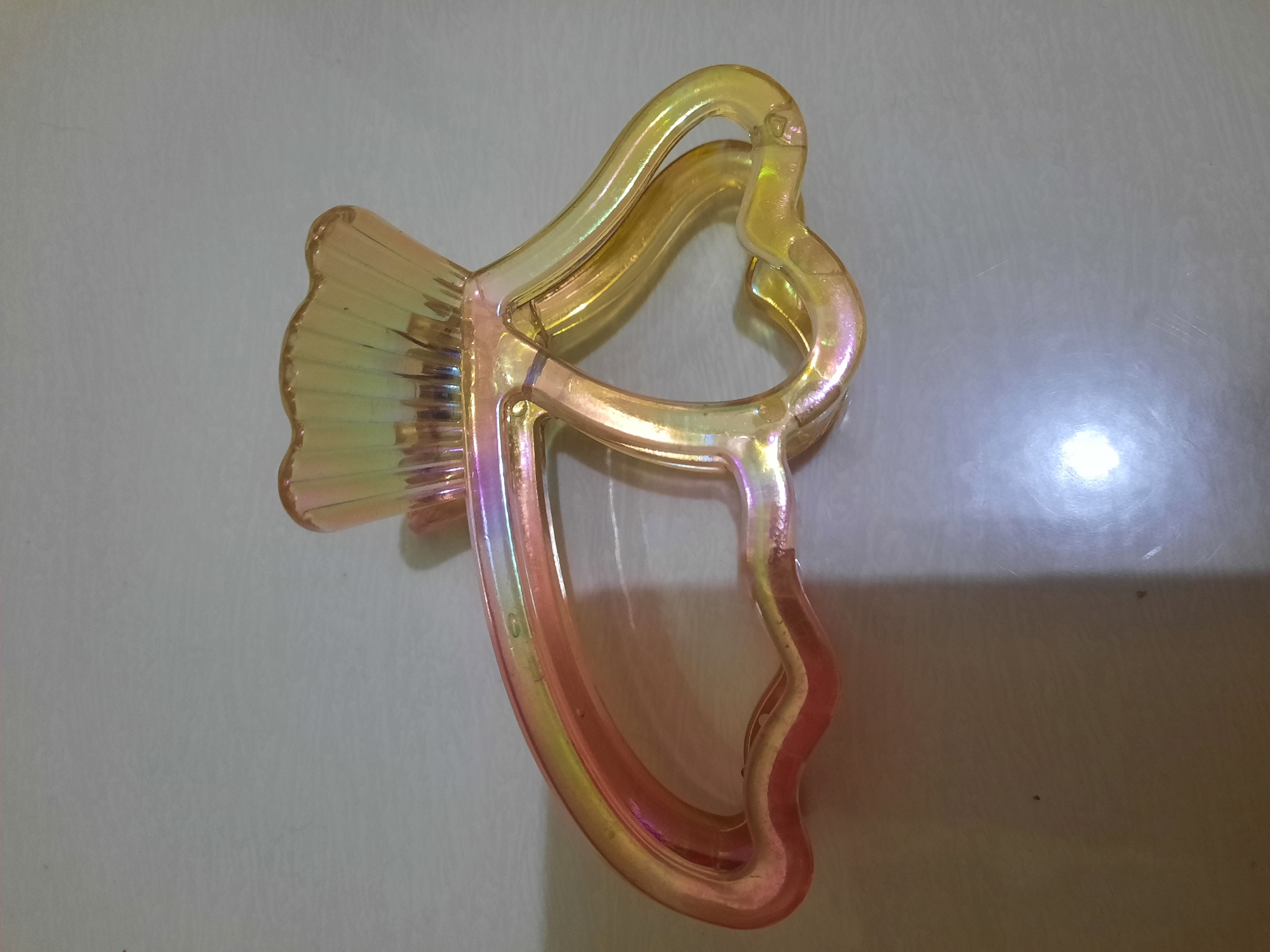 The image displays a single iridescent hair claw. The claw is made of a translucent plastic material that shimmers with shades of pink, yellow, and green. It has a curved, shell-like shape with a decorative, scalloped edge at the top. The hair claw is lying flat on a light-colored, textured surface, possibly a tabletop or countertop. The lighting in the image creates a bright reflection on the surface below the object. There is no visible indication of the city of Lhokseumawe, Indonesia.