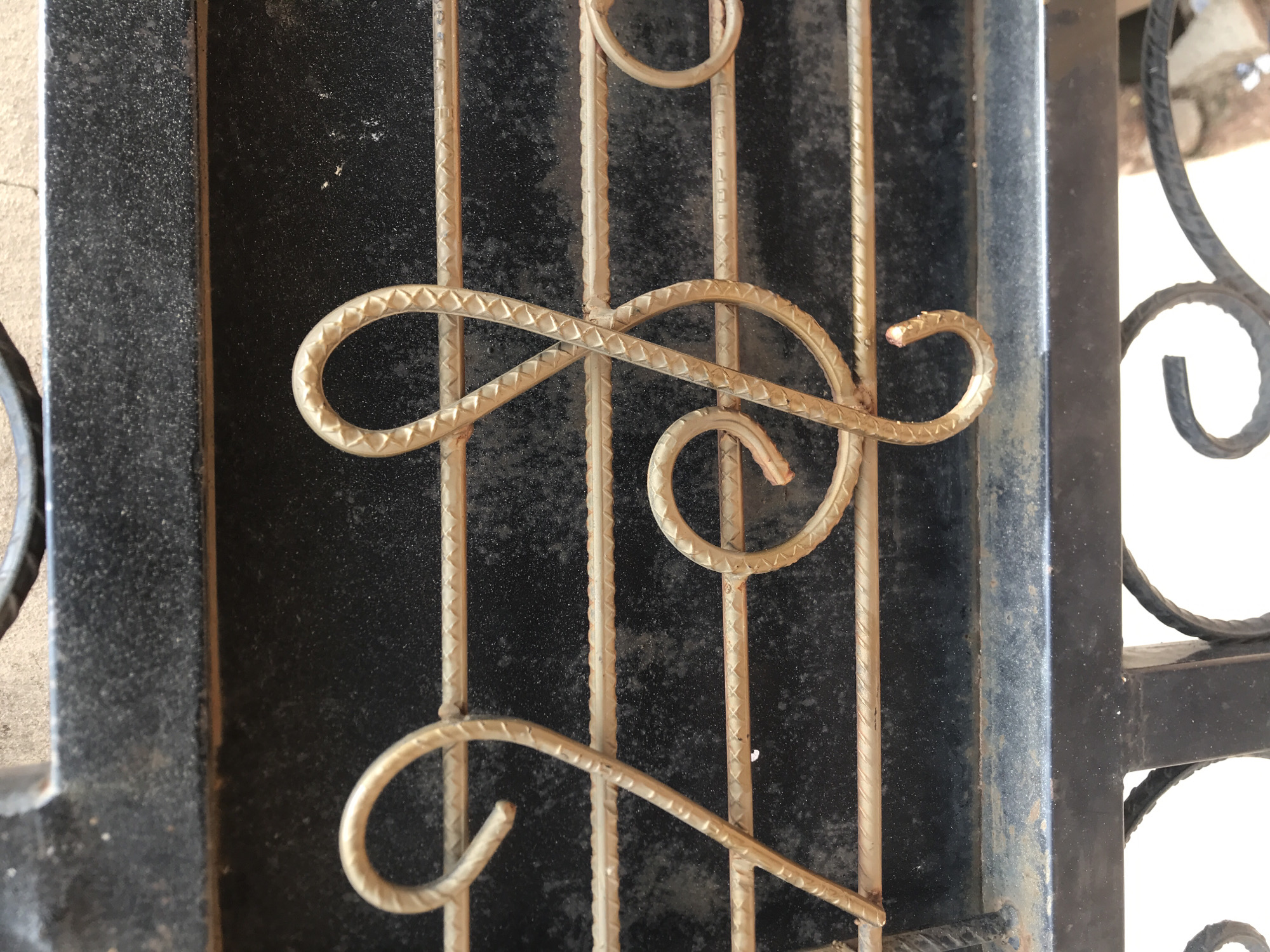 A detailed, eye-level photograph captures a segment of a metal gate or railing in Jos, Nigeria. The scene features five horizontal bars, constructed from golden-bronze reinforcing bar (rebar) with a distinct, repeated diamond-shaped knurling pattern. These bars are arranged parallel, forming a visual representation of a musical staff. On the left, a rebar element is shaped into a figure '2', while on the right, a rebar element forms a treble clef symbol. Both decorative elements match the material and finish of the horizontal bars. The background consists of a dark, granular-textured metal panel, appearing black. Visible above and below the rebar construction are wider, flat sections of black-painted metal framing. The metallic surfaces show minor surface wear and dust accumulation.