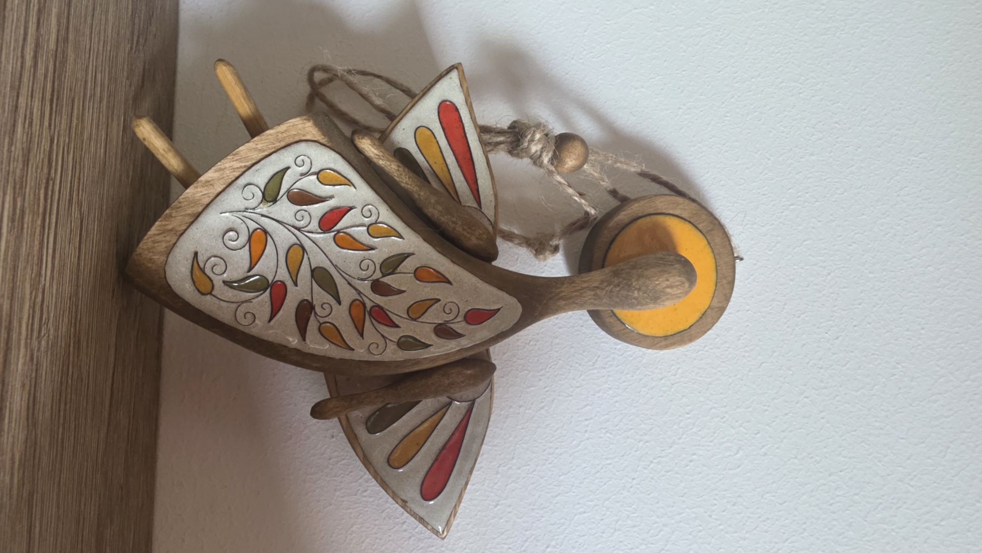 A wooden toy bird, possibly an ornament, is affixed to a light-colored wall. The bird is constructed from wood with painted decorations. Its body is roughly triangular, with leaf-like patterns in shades of orange, red, and green on a cream-colored background. It features wooden wings adorned with similar color schemes. A circular wooden piece with a yellow center is attached to the head, and a string with a wooden bead is used for hanging. The left edge of the frame shows a light brown wooden surface, likely a wall panel or furniture edge. The scene is not located in Saint Petersburg, Russia.