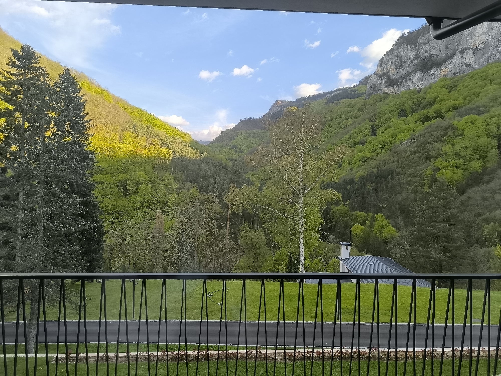 View from a balcony overlooking a mountainous landscape near Нареченски бани, Bulgaria. In the foreground, a black metal railing partially obscures a lawn. Beyond the lawn, a paved road divides the lawn from a forest area with a house featuring a gray roof and a white chimney. The background features forested mountains under a partially cloudy sky. The forests exhibit varying shades of green. A cliff face is visible on the right. The view is framed by a building structure above the balcony.
