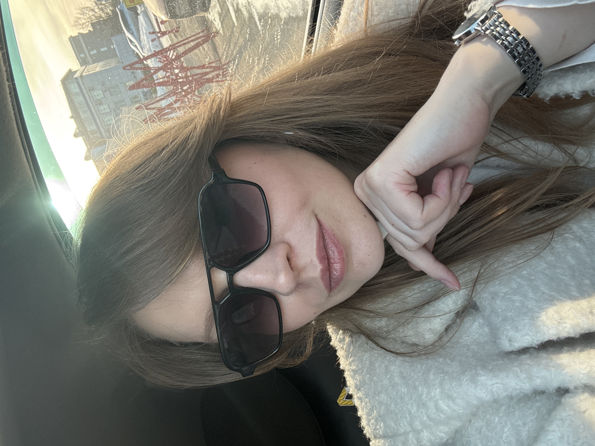 The scene depicts a selfie taken inside a vehicle in Izhevsk, Russia, on a sunny winter day. A female subject occupies the foreground, looking towards the camera. She has long, light brown hair and wears black-framed aviator-style sunglasses and a cream-colored, textured coat. Her right hand is positioned with fingers resting lightly near her chin. A silver-toned watch with a link bracelet is visible on her left wrist. The vehicle's dark interior is visible on the left and top. Through the window, an urban winter landscape is visible, characterized by strong sunlight and glare. Snow covers the ground. In the mid-ground, multiple red, geometric, possibly decorative metal structures are present, partially obscured by snow. Beyond these structures, multi-story buildings and other parked vehicles are discernible. Bare trees are visible in the distant background.