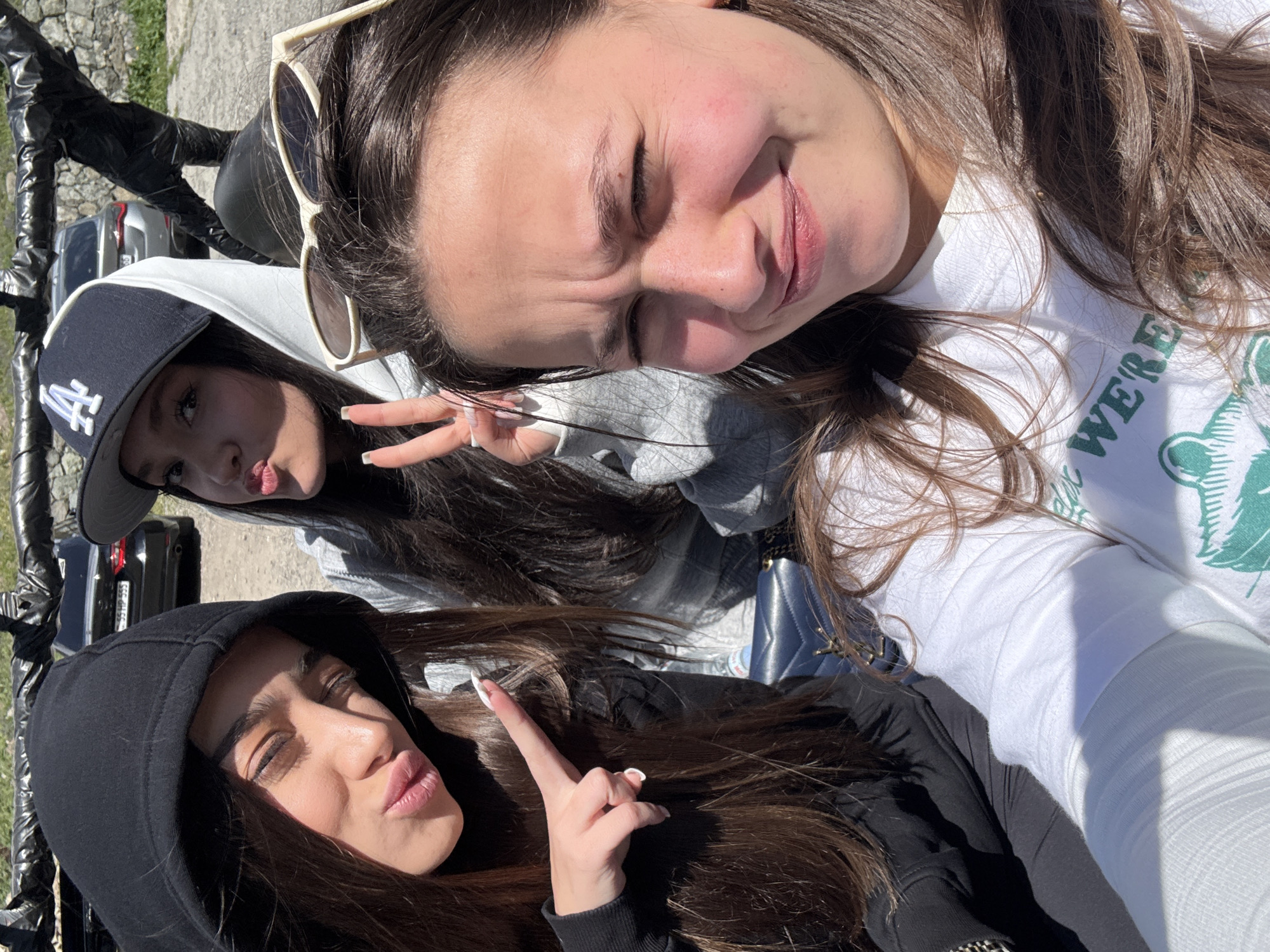 Three young women are visible in a selfie-style photograph. The woman at the top right is wearing a white T-shirt and has sunglasses perched on her head. She is smiling with her eyes closed. To her left and slightly below is a woman in a gray hooded sweatshirt and an "LA" baseball cap, making a peace sign. Below her is another woman wearing a black hooded sweatshirt, also making a hand gesture with a pursed-lip expression. Behind them is a black vehicle with a partial license plate visible, reading "55 HP 555." The location appears to be outdoors, with a rough stone or concrete surface visible in the background. Given the prompt's information, the location is likely Teghut, Armenia.