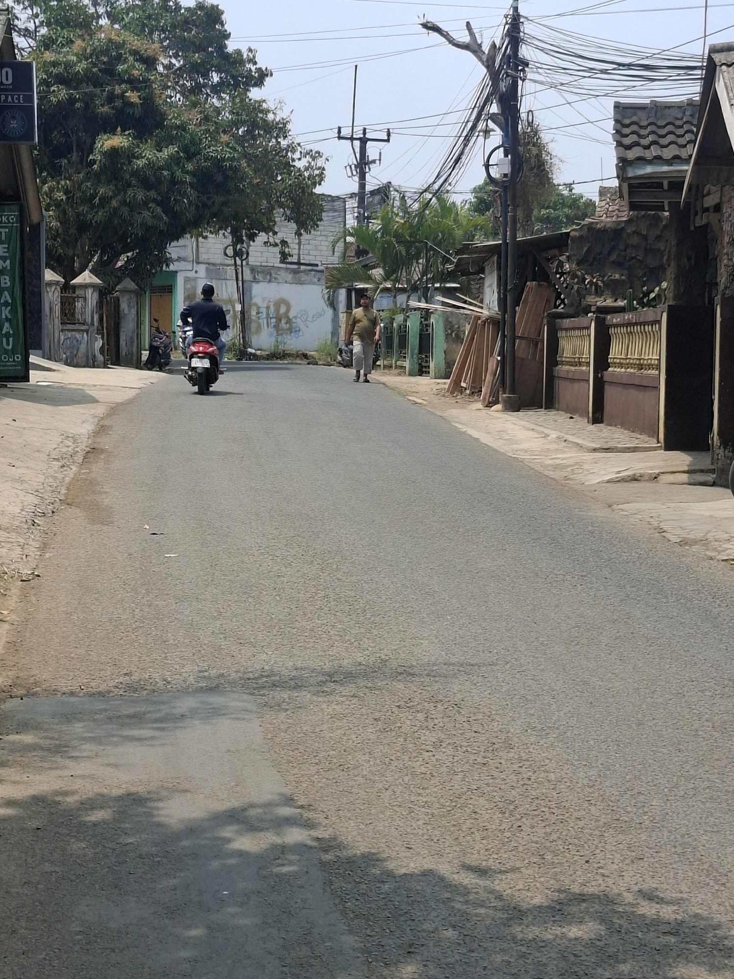 The image depicts a street scene in Ciheulang Tonggoh, Indonesia. A paved road occupies the majority of the frame, with a slight upward slope. On the left side of the road, there is a sidewalk with a concrete edging and some debris. Adjacent to the sidewalk are houses and buildings with varying architectural styles, some featuring fences and gates. A prominent feature on the left is a tall, dark pole with numerous electrical wires. In the middle ground, a person on a red motorcycle is riding away from the camera. Further down the road, another person is walking. The environment appears to be a residential or mixed-use area with trees and vegetation visible, particularly on the left side of the road. The sky is clear and bright, suggesting daylight. Shadows cast by trees are visible on the road, indicating the time of day.