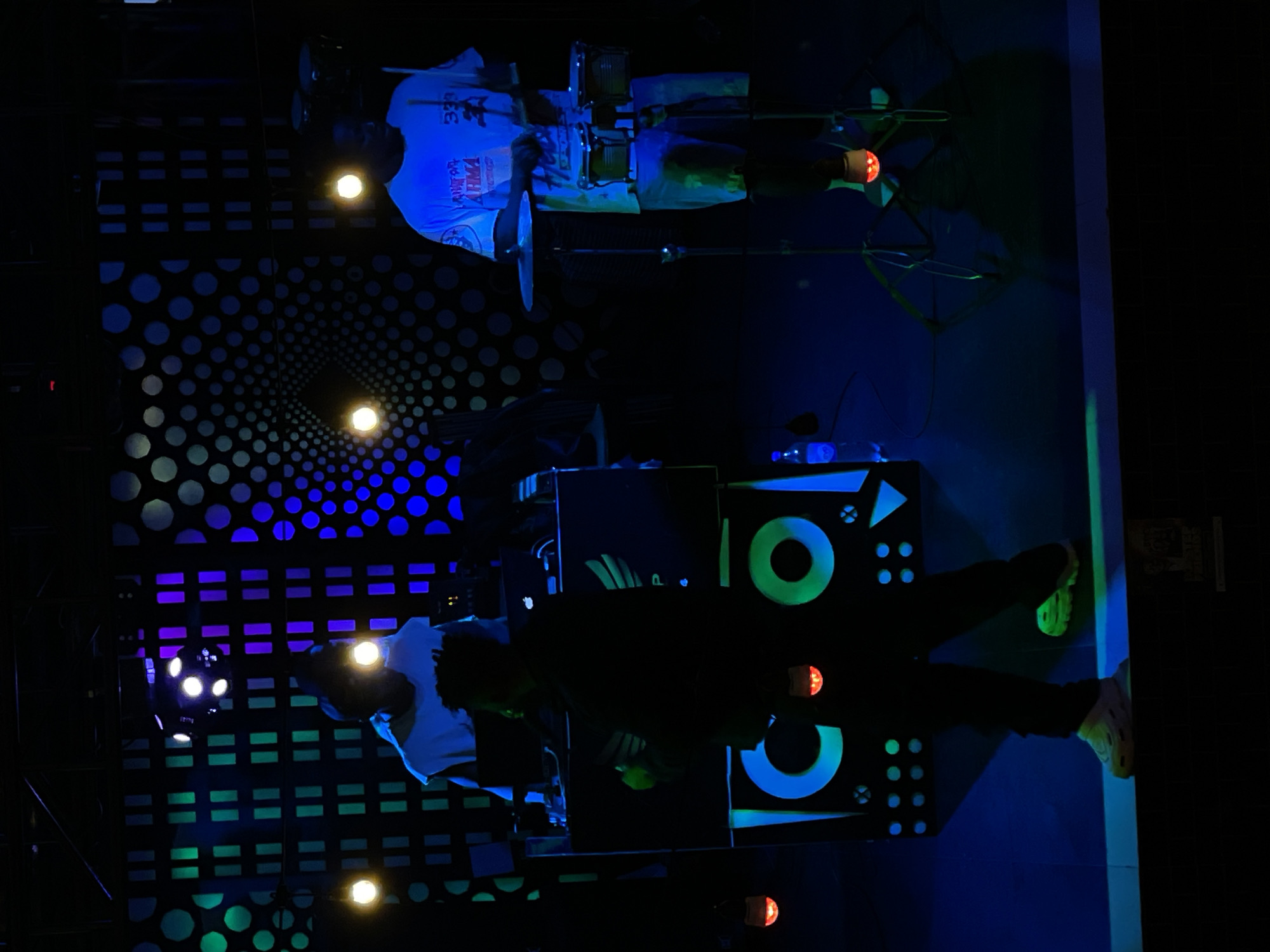 The image depicts an indoor scene with performers on stage, likely in a venue located in Obosi, Nigeria. The background features geometric light patterns, including circles and rectangles illuminated in blue, green, and purple. Two individuals are visible, one appearing to play drums and another at a DJ setup. The drummer wears a light-colored shirt with lettering on it. The DJ setup has blue and green accents. Lighting fixtures are scattered throughout, creating spots of illumination.