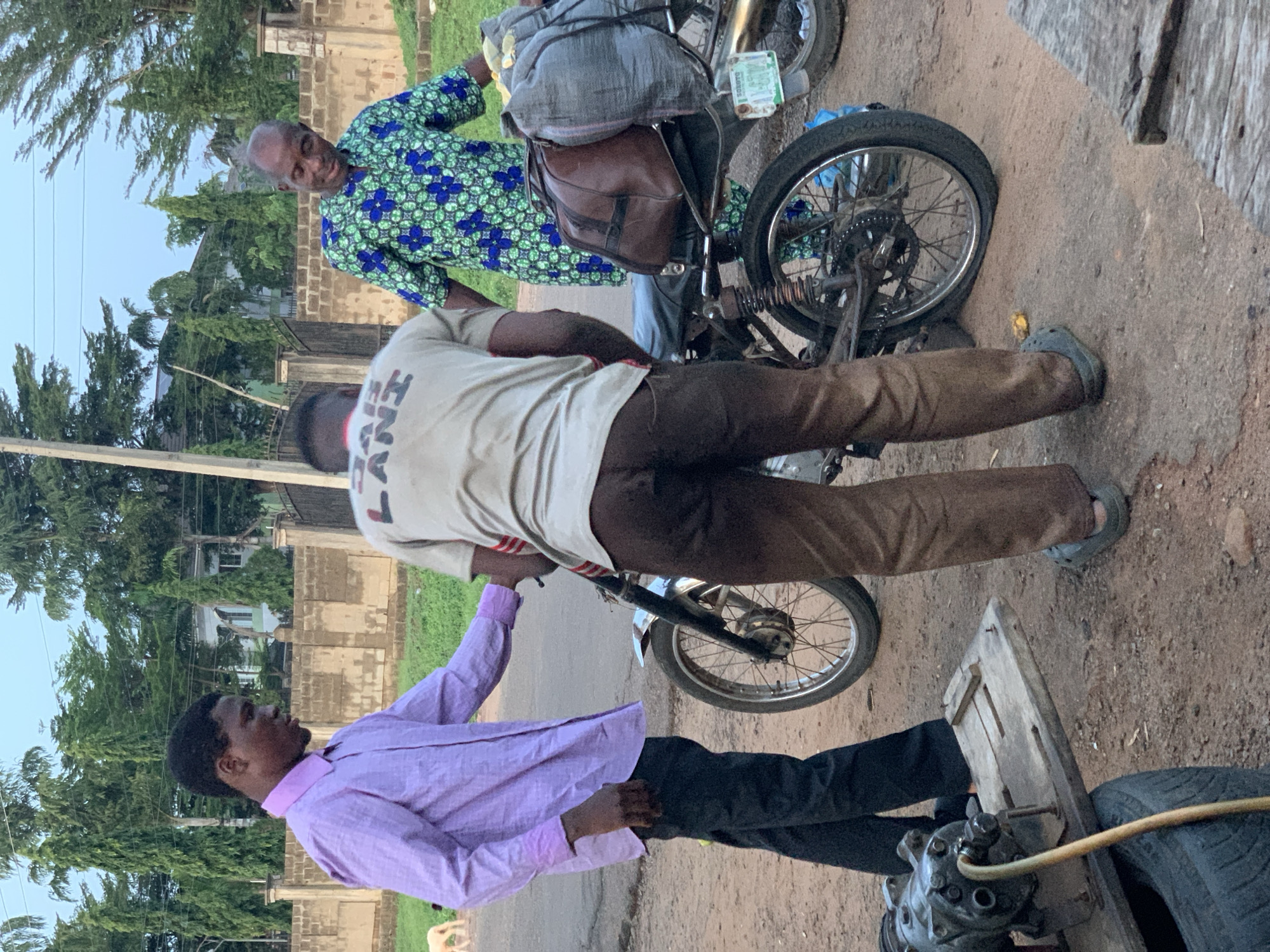 The image shows three men and a motorcycle in Gbeleko, Nigeria. One man is wearing a purple long-sleeved shirt and black pants. Another man is wearing a tan t-shirt and brown pants, and the third man is wearing a green and blue patterned shirt. The men are standing near a motorcycle which is loaded with luggage and a license plate. An air compressor sits on the ground by a spare tire. The surrounding environment shows a paved road and trees.