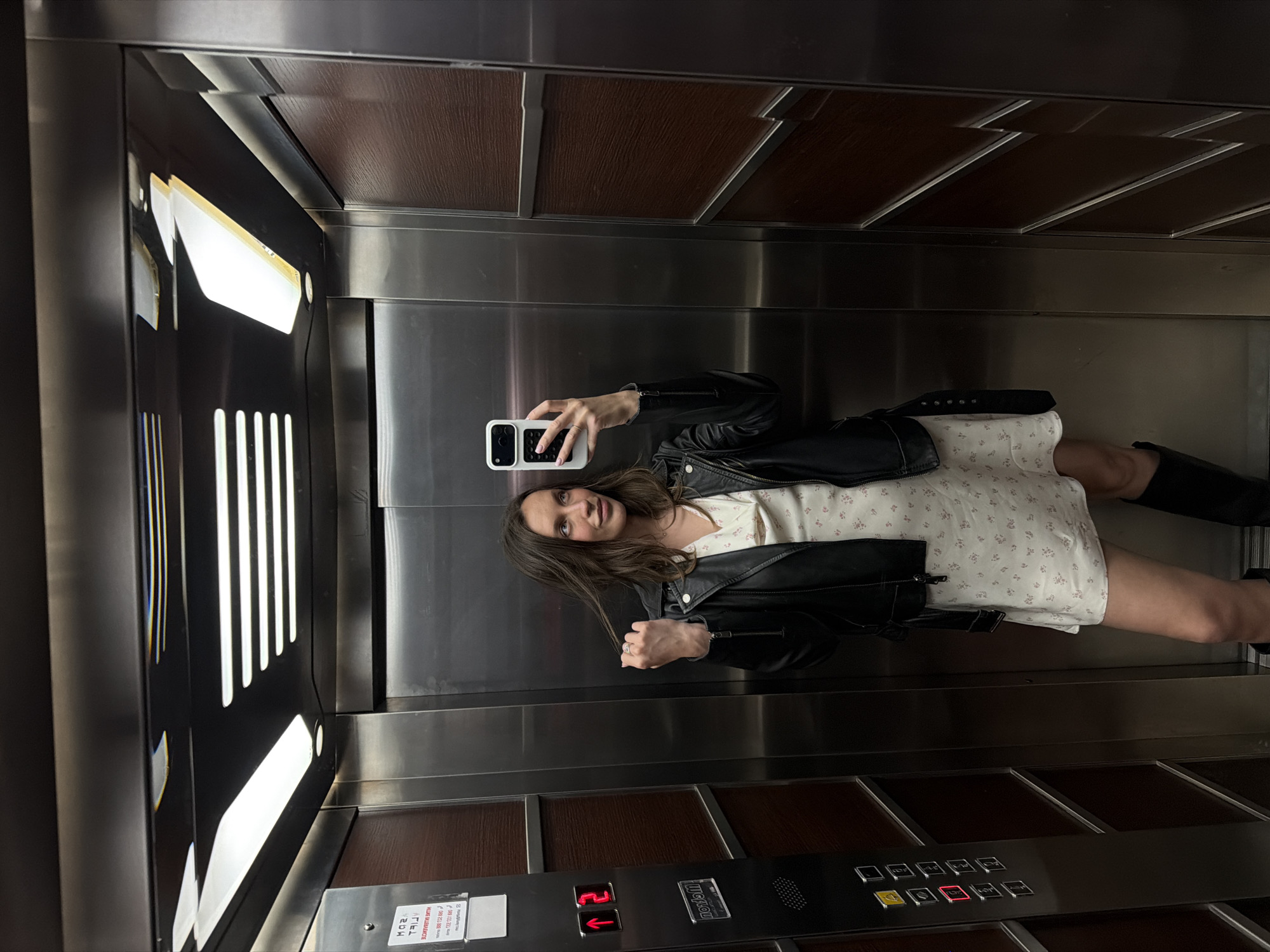 The image, rotated 90 degrees counter-clockwise, depicts a young woman taking a mirror selfie inside an elevator. She is positioned centrally, holding a white smartphone with a black attachment in her right hand, which obscures part of her face. She has long, dark brown hair and a faint smile. She is dressed in a black leather jacket over a light-colored, short-sleeved dress with a subtle floral pattern, paired with dark boots.
The elevator interior features reflective stainless steel walls, which serve as the mirror for the selfie. Dark brown wooden panels, segmented by metal frames, are visible above and below the main mirrored section. Linear light fixtures are present on the left wall of the elevator.
On the lower right, an elevator control panel displays the illuminated number "2" and a red up-arrow, indicating the current floor and direction. The brand name "Rosan" is visible on the panel, alongside several square buttons. A small, mirrored sticker in the bottom left corner shows partial text, including what appears to be contact numbers like "06X XXX XX XX" and "888 722 00". The setting is an indoor space within Budva, Montenegro.