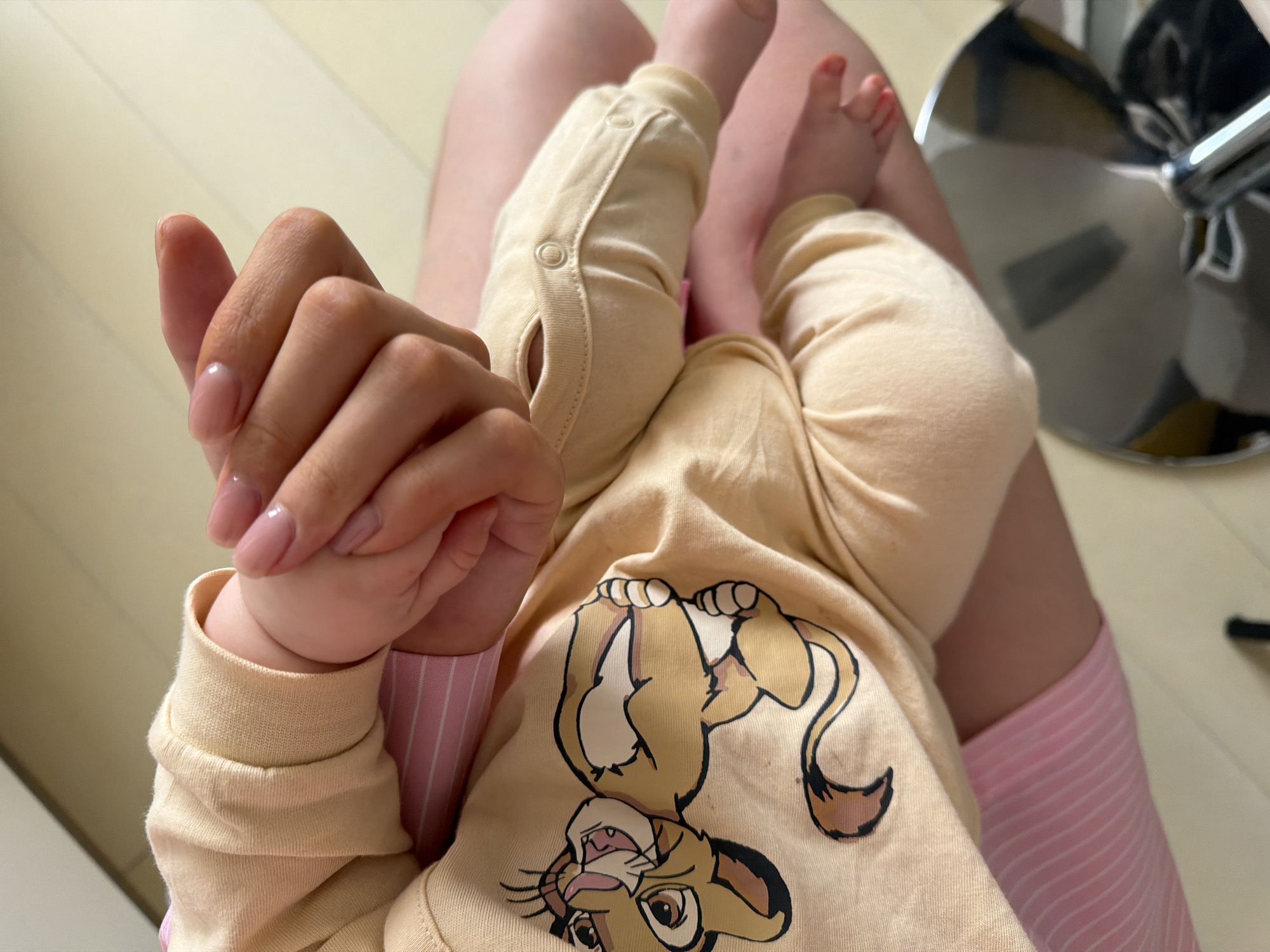 The image shows a person holding a baby dressed in a beige outfit with a Simba (The Lion King) graphic. The baby is lying on the person's lap, with the baby's legs bent and feet visible. The person holding the baby has pink fingernails. The background includes the person's legs in pink striped shorts, a portion of a chrome office chair, and a light-colored wooden floor. There is no information available about the location in Khimki, Russia.