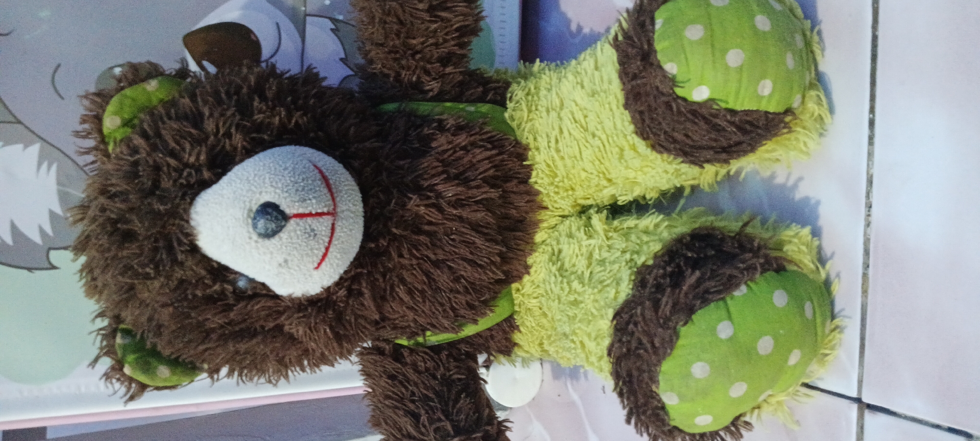 This image features a close-up, top-down view of a fluffy, stuffed teddy bear lying on a light-colored, tiled floor, possibly in a home setting in Rejing, Indonesia. The bear has brown, shaggy fur and is adorned with lime green patches featuring white polka dots on its paws and ears. Its face is white with a blue eye, a red stitched mouth, and a red stitched nose. The bear's body is segmented into alternating bands of brown fur and lime green fuzzy material. The lighting suggests it is daytime, and the shadows indicate artificial light is also present. No people are visible, and the primary focus is the solitary stuffed toy.