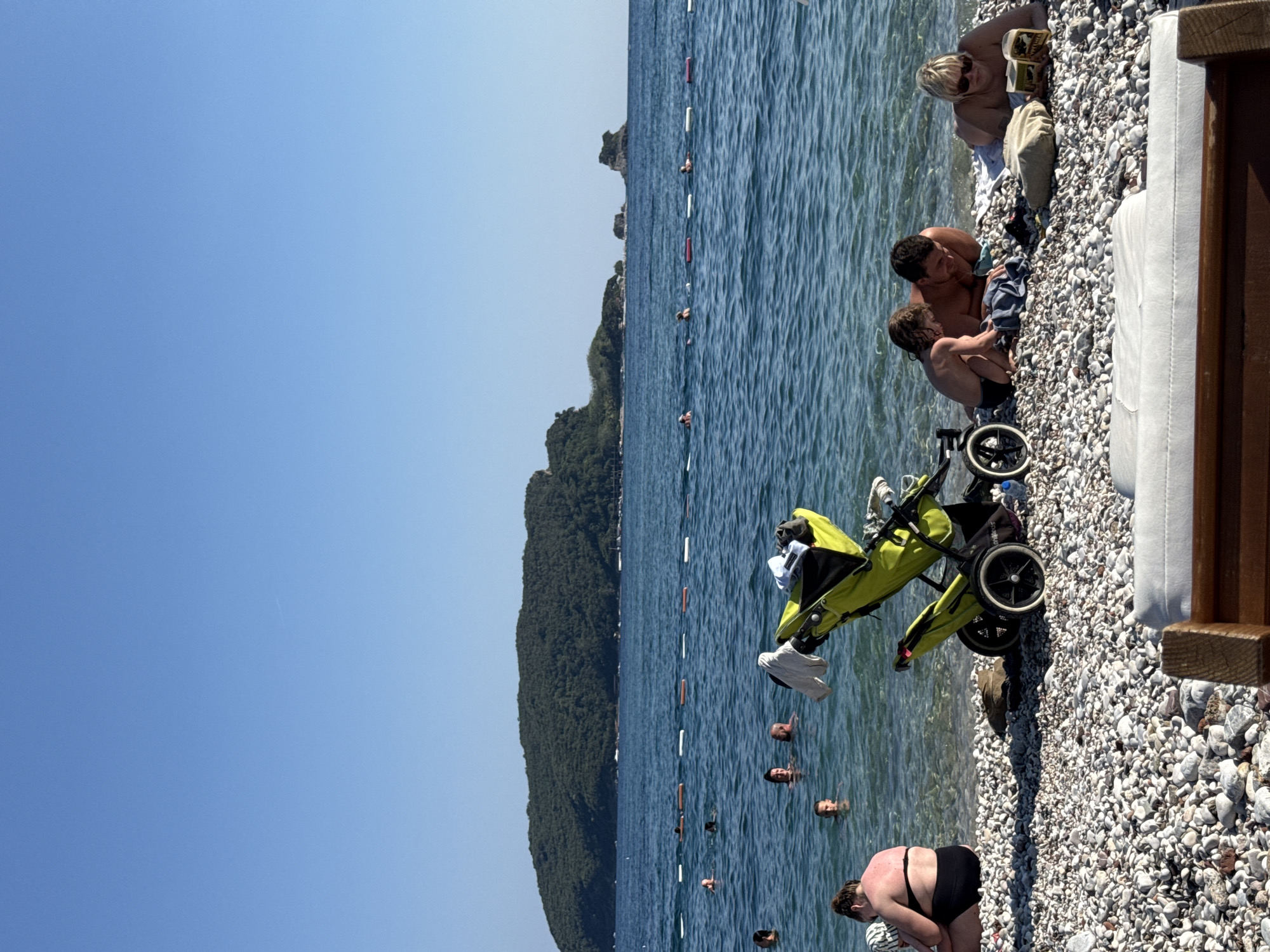 The image depicts a sunny day at a pebble beach in Budva, Montenegro. The scene is divided between the water and the shore, with a clear blue sky above. In the water, several people are visible, some swimming and others wading. A line of buoys marks a designated swimming area. Lush green hills rise in the background, typical of the Montenegrin coast.
On the shore, a family is gathered near the water. A man and a young child are interacting, while another woman, possibly reading, is seated nearby. A bright green stroller with large wheels is positioned on the pebble beach, suggesting it's ready for use or has just been used. The sunlight casts shadows on the pebbles, indicating it's likely midday. The overall atmosphere appears relaxed and leisurely, with people enjoying a day at the beach. No visible text is present in the image.