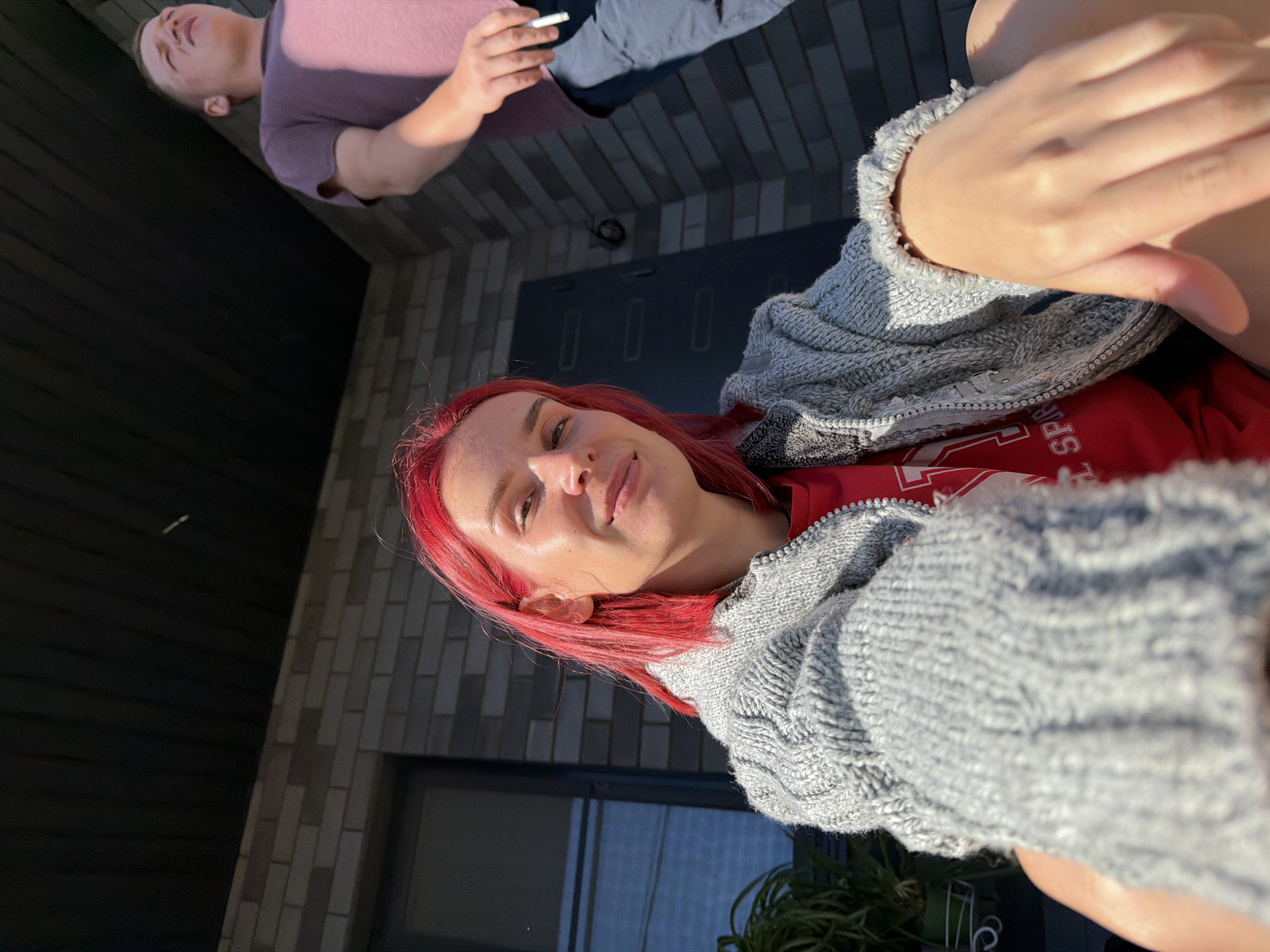 A low-angle, close-up selfie shows a woman with bright red hair, wearing a gray knitted cardigan over a red t-shirt, looking directly at the camera. Her left arm is in the foreground. Behind her, to the right and slightly out of focus, a man in a purple t-shirt and dark pants stands, holding a cigarette. They appear to be on the porch of a brick house. The environment suggests an outdoor setting, possibly a suburban area of Chaikovsky, Russia, under a bright, sunny sky, indicated by the strong shadows and sunlight.