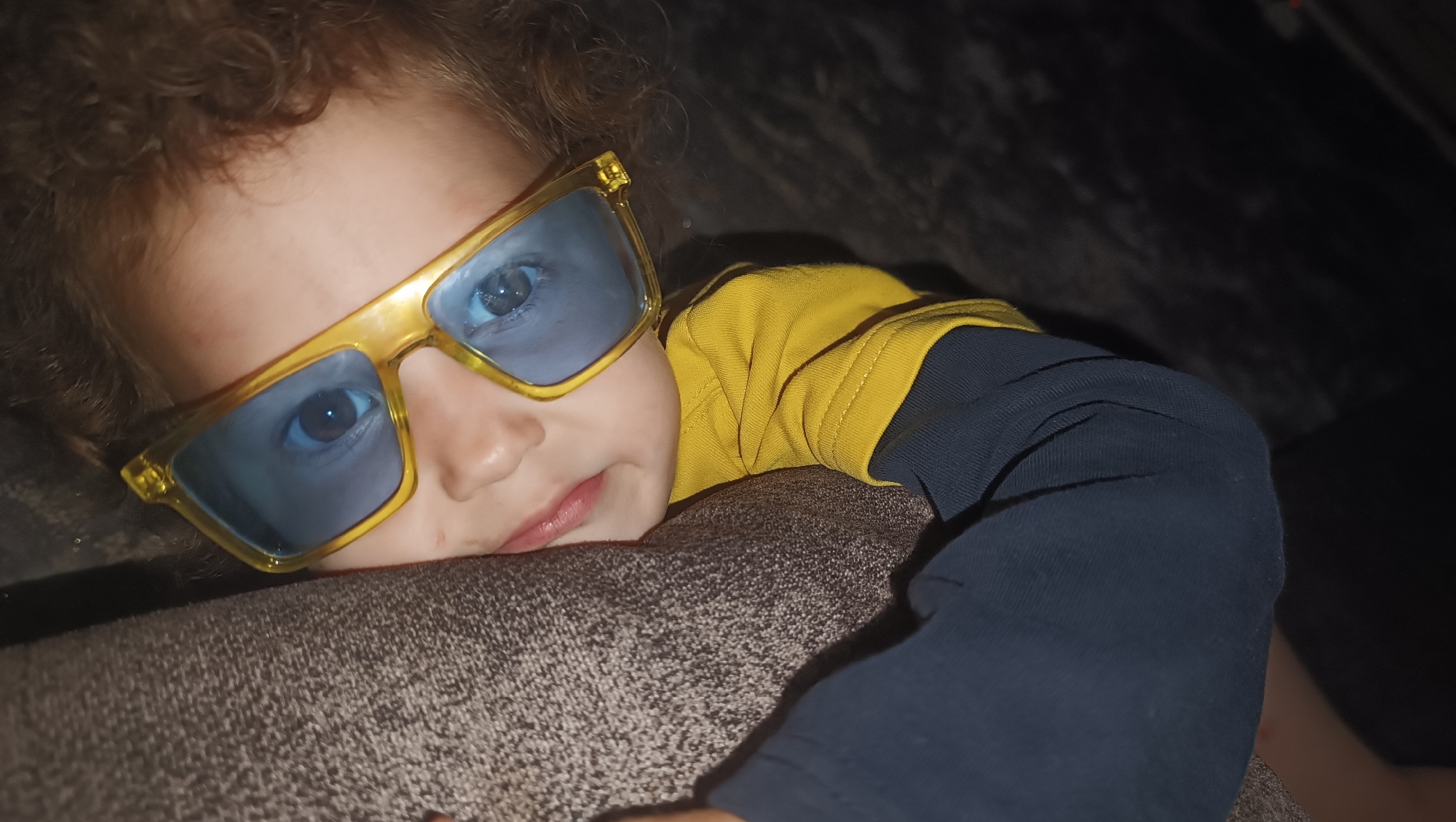 A close-up, low-angle shot shows a young child lying on a textured, brown surface, possibly a sofa. The child has curly brown hair and is wearing large, yellow-framed glasses with blue lenses. They are also wearing a yellow long-sleeved shirt with navy blue sleeves. The child's head is resting on the brown surface, and their gaze is directed towards the camera. The lighting is dim, creating a dark background. The image does not contain information about the city of Karsha, Russia.