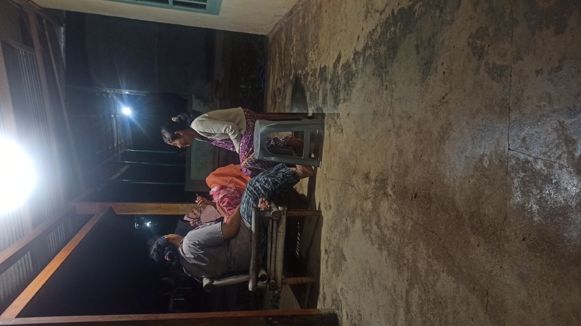 The image shows three people, appearing to be Indonesian women, conversing on a porch at night. The woman on the left is seated on a bamboo bench and wearing a grey t-shirt and patterned pants. The woman in the middle is lying on a pink and orange blanket. The woman on the right is sitting on a grey plastic stool and wearing a light-colored cardigan over a patterned top and skirt. The porch has a corrugated metal roof supported by wooden beams. The floor is made of concrete with visible cracks and water stains. A bright light source, likely a bulb, illuminates the scene from the roof. In the background, there are dark shapes suggesting trees or other structures. The overall impression is of a casual, outdoor gathering in a rural or semi-rural setting, possibly in the city of Tuba, Indonesia, given the context.