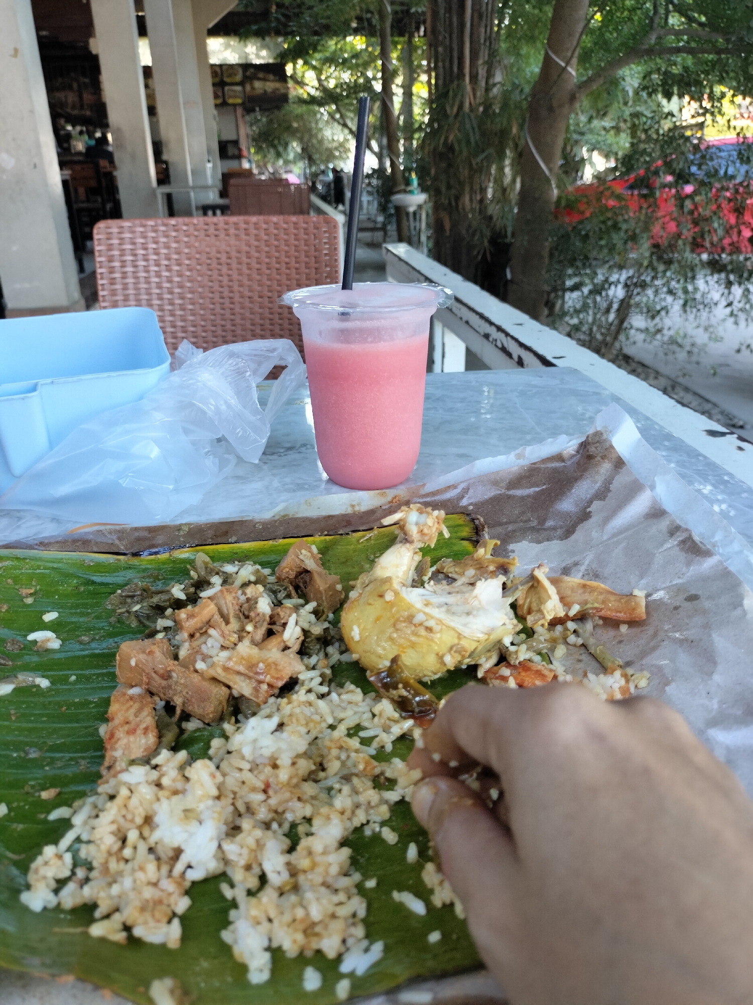 The image depicts an outdoor dining scene at Keudealue Ie Puteh, Indonesia, featuring a meal served on a banana leaf. In the foreground, a portion of chicken and rice is spread across the leaf, with a hand reaching in to pick up food. Beside the meal is a pink beverage in a clear plastic cup with a straw. A light blue plastic container and a crumpled plastic bag are also visible on the table. The background shows a covered seating area with rattan chairs and tables, suggesting a restaurant or food stall. Trees and other vegetation can be seen beyond the seating area, indicating an outdoor setting. The lighting suggests it is daytime.