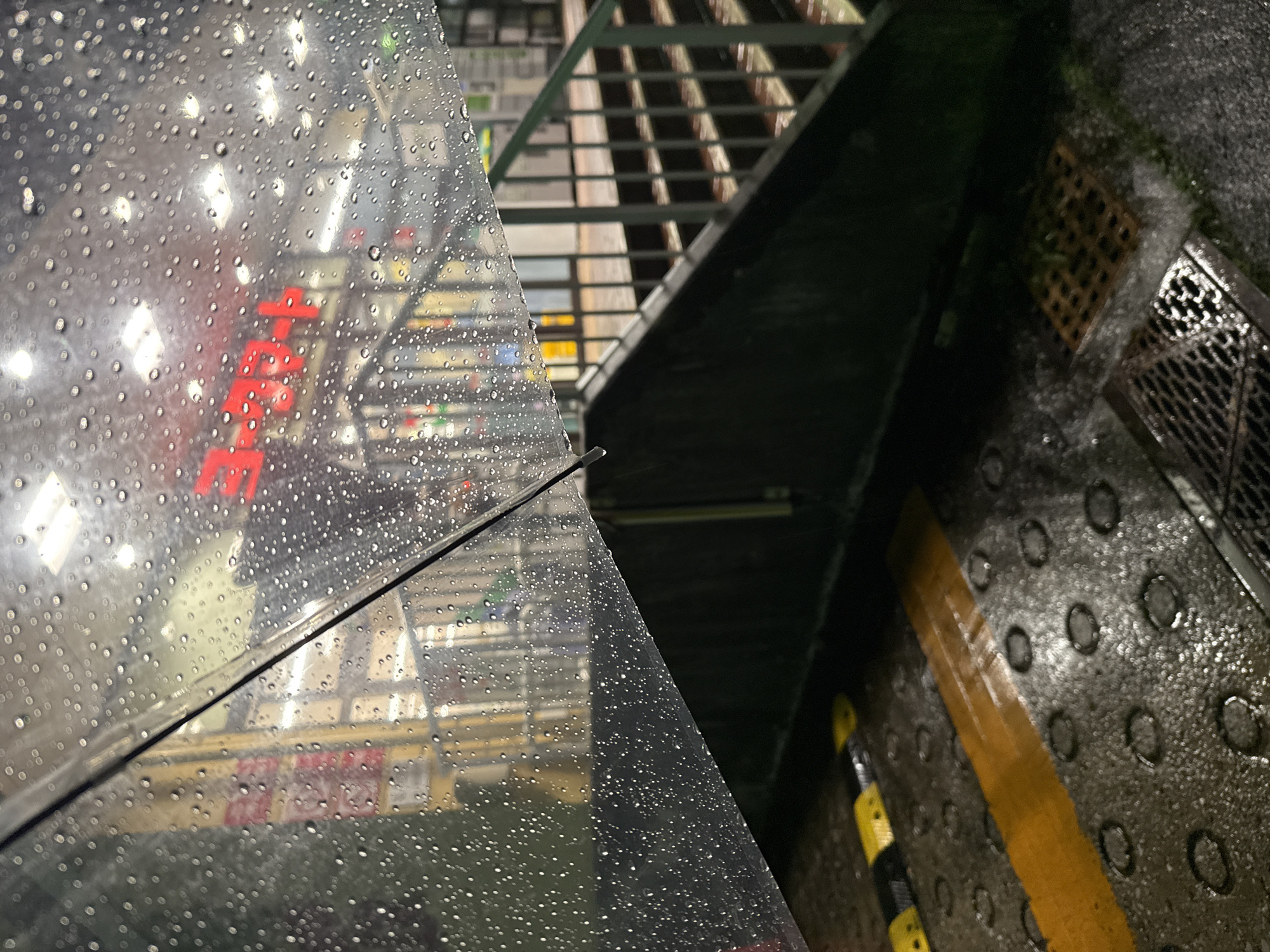 The image captures a wet outdoor scene, likely at a train station or a similar public transportation hub in Yachiyo, Japan. The foreground is dominated by a transparent umbrella speckled with raindrops, partially obscuring the view. Below the umbrella, the ground is wet and reflective, with yellow and black hazard markings and circular indentations visible. A metal grate is also present on the wet surface. In the background, a set of stairs with metal railings leads upwards. Above the stairs, a red digital display, possibly a clock or arrival board, is illuminated. The environment appears dimly lit, suggesting it is nighttime or a heavily overcast day, and the presence of rain is evident throughout the scene. No people are discernible in the image.