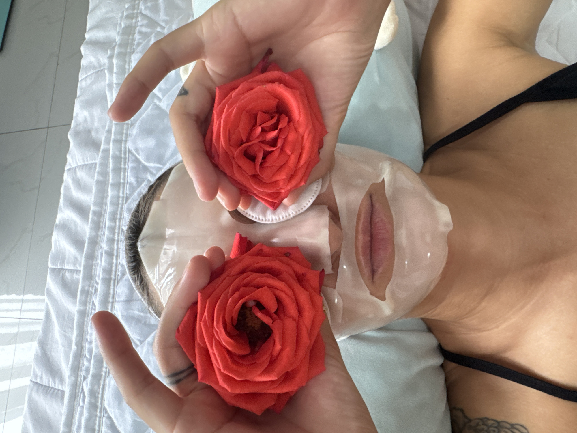 A person, lying down on a quilted surface, is holding two red roses up to their face. The person is wearing a white sheet face mask. One rose is held over their left eye, and the other rose is held over their right eye. The person's hands are visible, with tattoos on the fingers. The background consists of a white quilted surface and a light blue pillow. The scene appears to be indoors, possibly in a bedroom. There is no indication of the location being in Nha Trang, Vietnam.