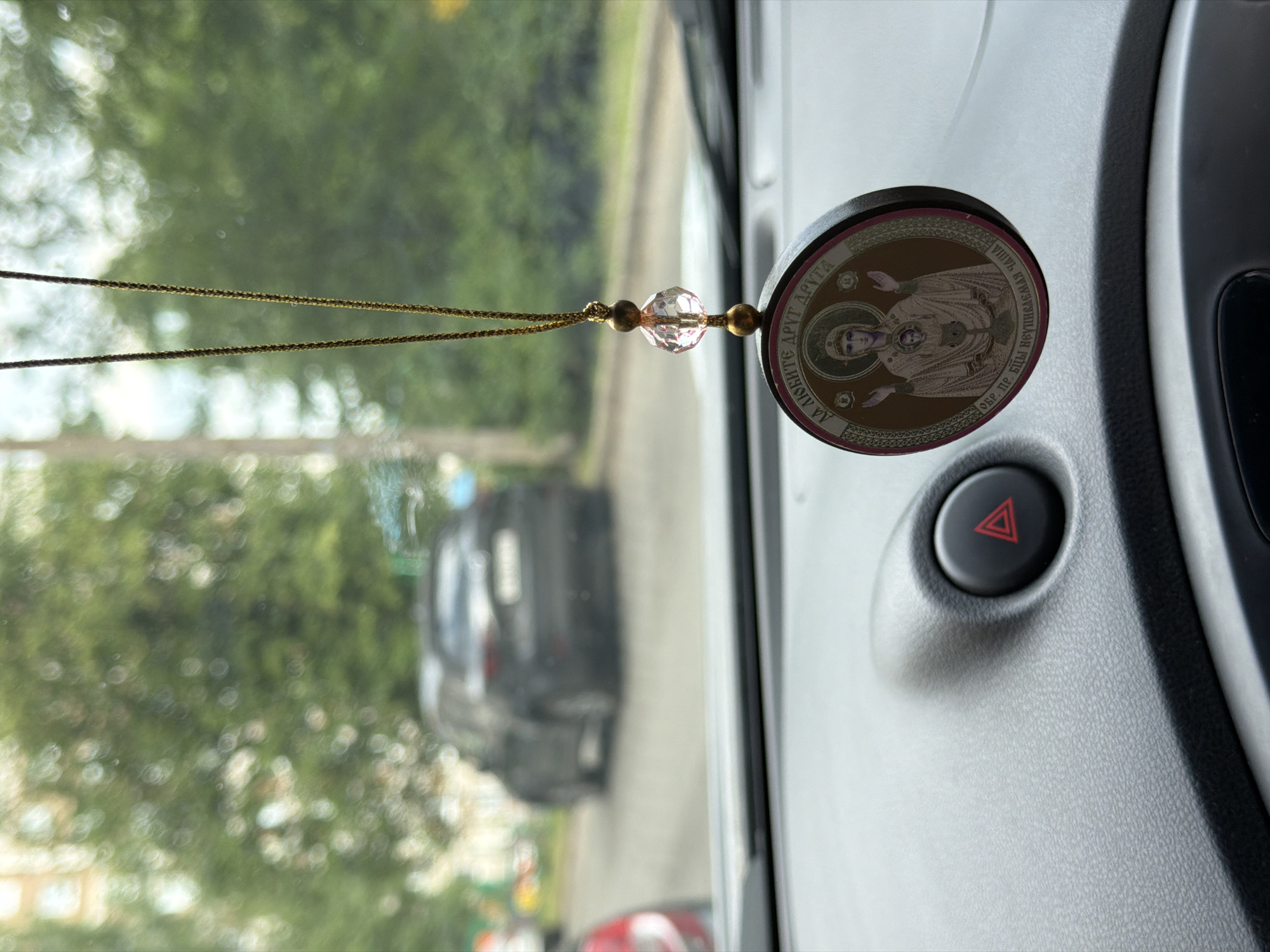 This image depicts a close-up view of a car's interior, specifically focusing on a religious ornament hanging from the rearview mirror. The ornament is circular and features an icon of the Virgin Mary holding the Christ child. In the background, through the windshield, a dark-colored SUV is visible on a street, suggesting an outdoor setting. Trees and foliage are blurred in the background, indicating a somewhat green environment, possibly a residential area or park. The dashboard of the car is visible in the foreground, with a red hazard light button prominent on the right side. The overall impression is one of a parked or slowly moving vehicle in an urban or suburban environment.