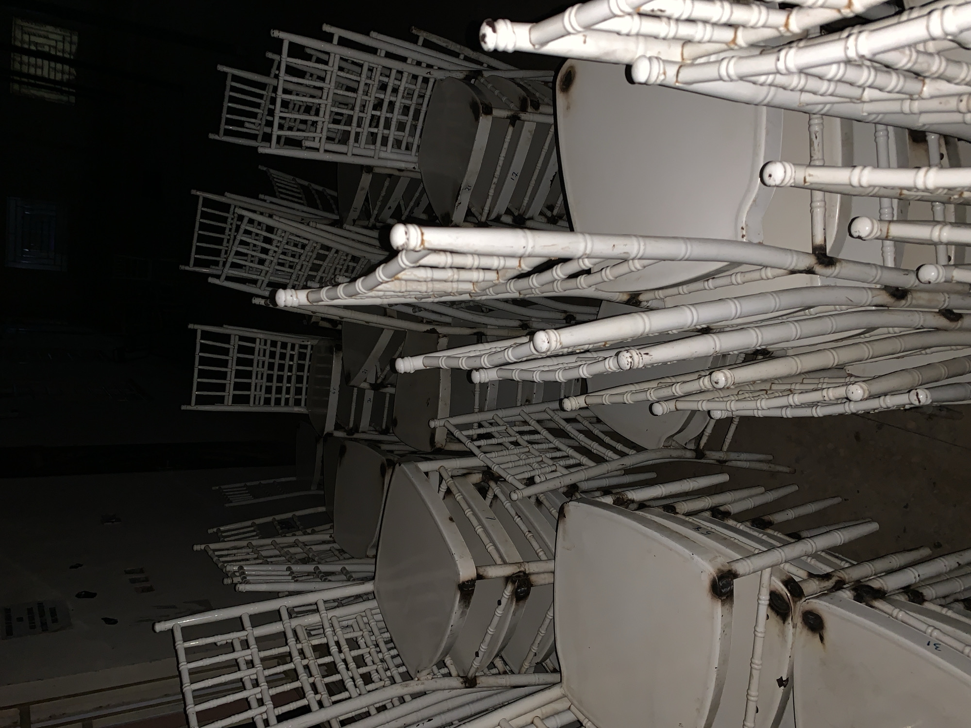 The image shows a stack of white, metal chairs with round seats in an indoor setting. The chairs are stacked closely together, occupying most of the frame. Some chairs show signs of wear and tear, including rust or discoloration at the welding points. The lighting is dim, casting shadows and obscuring the details of the room, although a wall and a window are discernible. The location is unknown. There are no people visible in the scene.