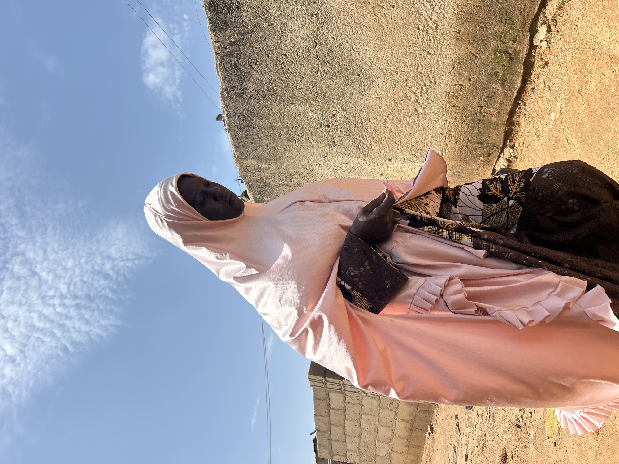 A young woman walks outdoors in the city of Jos, Nigeria, under a clear blue sky with scattered white clouds. She is wearing a light pink hijab that covers her head and shoulders, and a colorful patterned dress underneath. The woman is walking to the right of the frame, looking down. Her hands are clasped together in front of her. Behind her, a rough, beige-colored wall runs horizontally across the frame. To the left of the wall, a fence made of concrete blocks is visible, along with some construction materials. The ground is covered in dirt and gravel, suggesting an unpaved area. The lighting indicates it is daytime, with the sun casting shadows.
