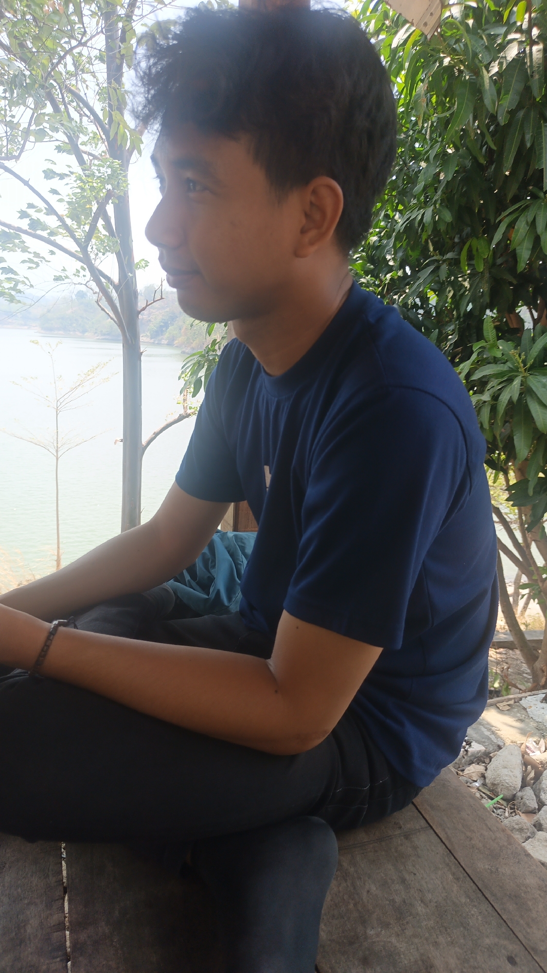 A young man with dark, curly hair is sitting in an outdoor setting, facing right and looking to the side. He is wearing a dark blue t-shirt and dark pants. His left arm is resting on his lap, and he is wearing a bracelet on his left wrist. The background features a body of water, trees with green leaves, and a wooden structure. The man appears to be relaxed, with his legs crossed. The scene is lit by natural sunlight.