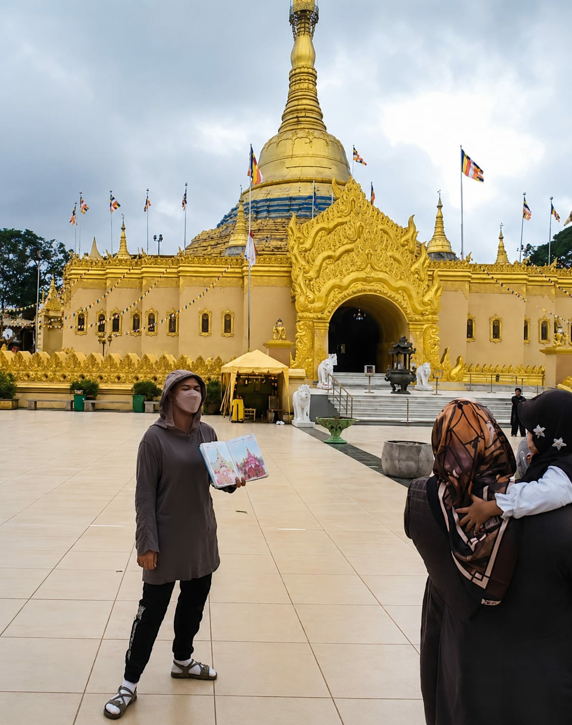 In Dolat Rayat, Indonesia, an outdoor scene unfolds in front of a large, ornate golden Buddhist temple. The temple features a prominent spire and intricate architectural details, with flags flying from poles in front of it. The foreground is paved with large square tiles, and the ground is mostly flat.
A woman stands in the center of the frame, facing slightly to the right. She is wearing a long, grey hooded garment, black pants, and white sandals. She has a light pink face mask covering her nose and mouth and is holding a book open in her hands, which displays images of colorful pagodas.
To the right of the woman, in the middle ground, an adult is carrying a young child on their shoulders. The adult is wearing a dark headscarf and a dark outer garment. The child, also wearing a headscarf, is looking towards the woman.
In the background, the main entrance to the temple is visible, with steps leading up to it. White lion statues flank the entrance. There are also some yellow tents set up to the left of the entrance. The sky above is overcast. The overall impression is one of a tourist or visitor engaging with the cultural and religious site.