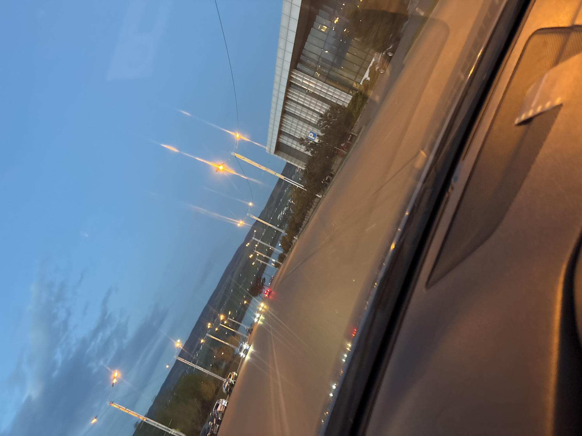 The media file presents an exterior scene captured from inside a vehicle at dusk or early night. A multi-lane road stretches through the frame, illuminated by numerous streetlights that emit a warm yellow-orange glow, causing prominent lens flares. Several cars, with illuminated headlights and red taillights, are visible on the road, suggesting movement.
To the right of the road, a large, modern building with a glass facade and many windows stands, its interior lights softly glowing. Near the base of this building, a blue "P" sign is visible, likely indicating a parking area. In the distance, beyond the road and partially obscured by trees, a body of water (possibly a river or lake) reflects scattered lights, with mountains or hills rising behind it.
The sky is a deep blue, indicative of twilight, with faint clouds visible in some areas. Reflections from the car's interior and dashboard are present on the windshield, framing the view. The overall impression is a dynamic, illuminated urban landscape during the transition from day to night.