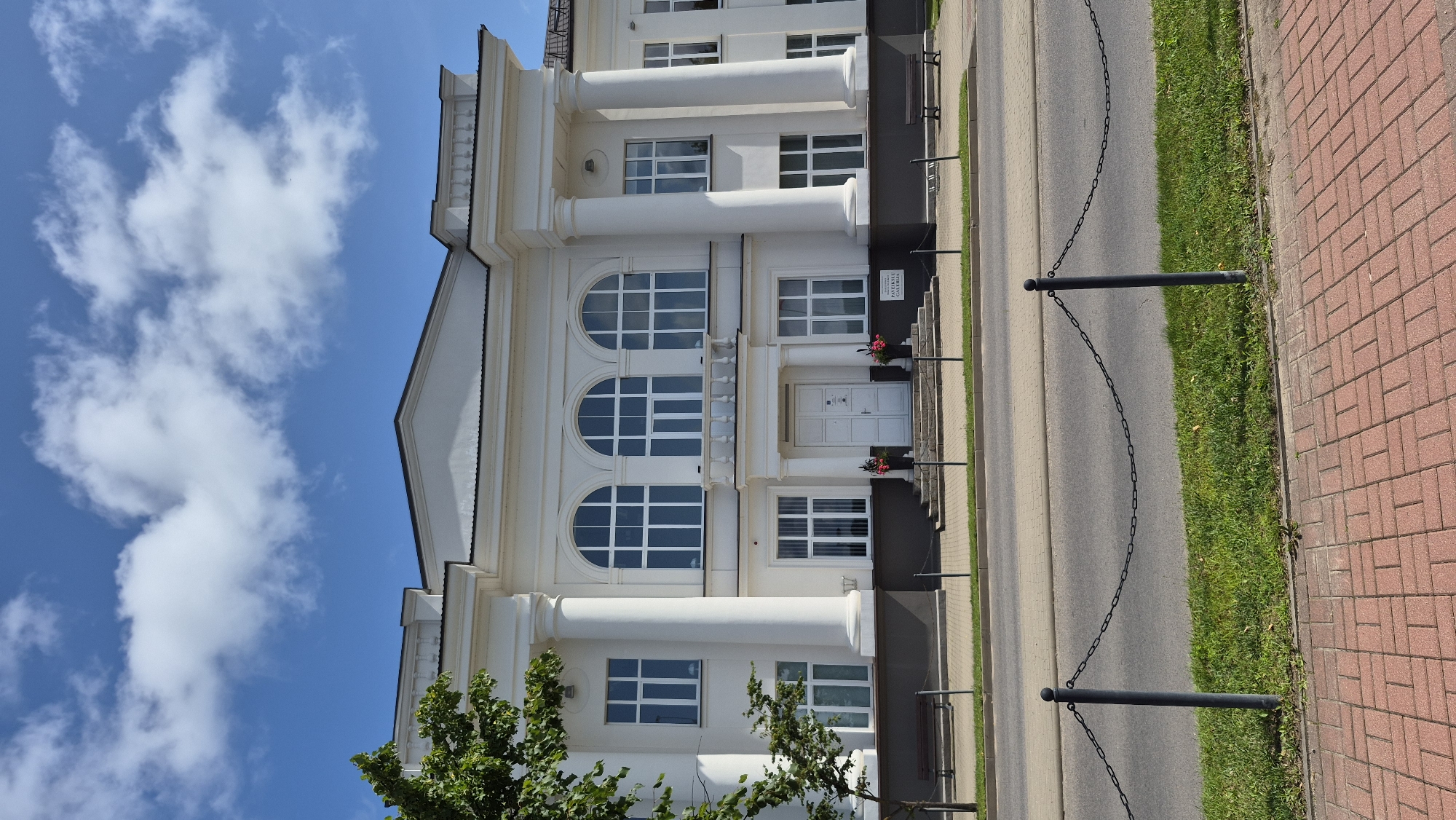 The image shows the facade of a white, neoclassical building in Zarasai, Lithuania. The building features a central entrance with double doors, flanked by tall columns and arched windows. A balcony with a balustrade is visible above the entrance, supported by columns. The building is situated across a street from a sidewalk and a grassy area, which is bordered by a chain-link fence. The sky above is blue with scattered white clouds. A tree with green leaves is partially visible on the left side of the image. Benches are placed along the sidewalk in front of the building. A sign with text, possibly indicating the name of the establishment, is visible on the right side of the entrance. The overall scene appears to be a public space, likely a cultural or administrative building, in Zarasai.