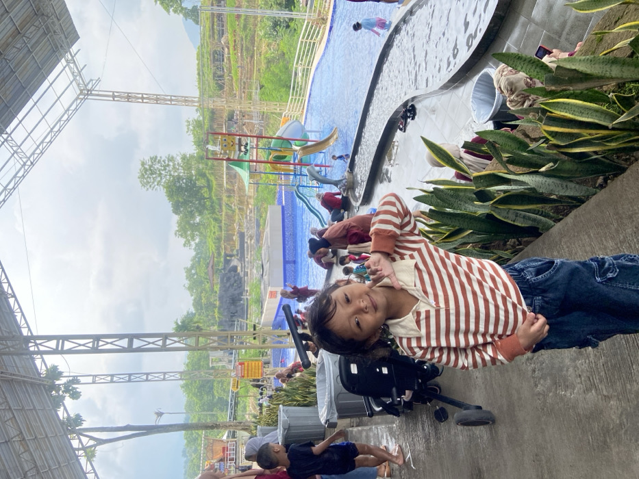 A young child in a striped shirt and jeans stands on a concrete surface near a plant bed, posing with a peace sign. Behind them is a water park with swimming pools and slides. Other people are in and around the pool area. A stroller sits nearby. Above, there are metal structures and some foliage on a hill. The sky is overcast.
