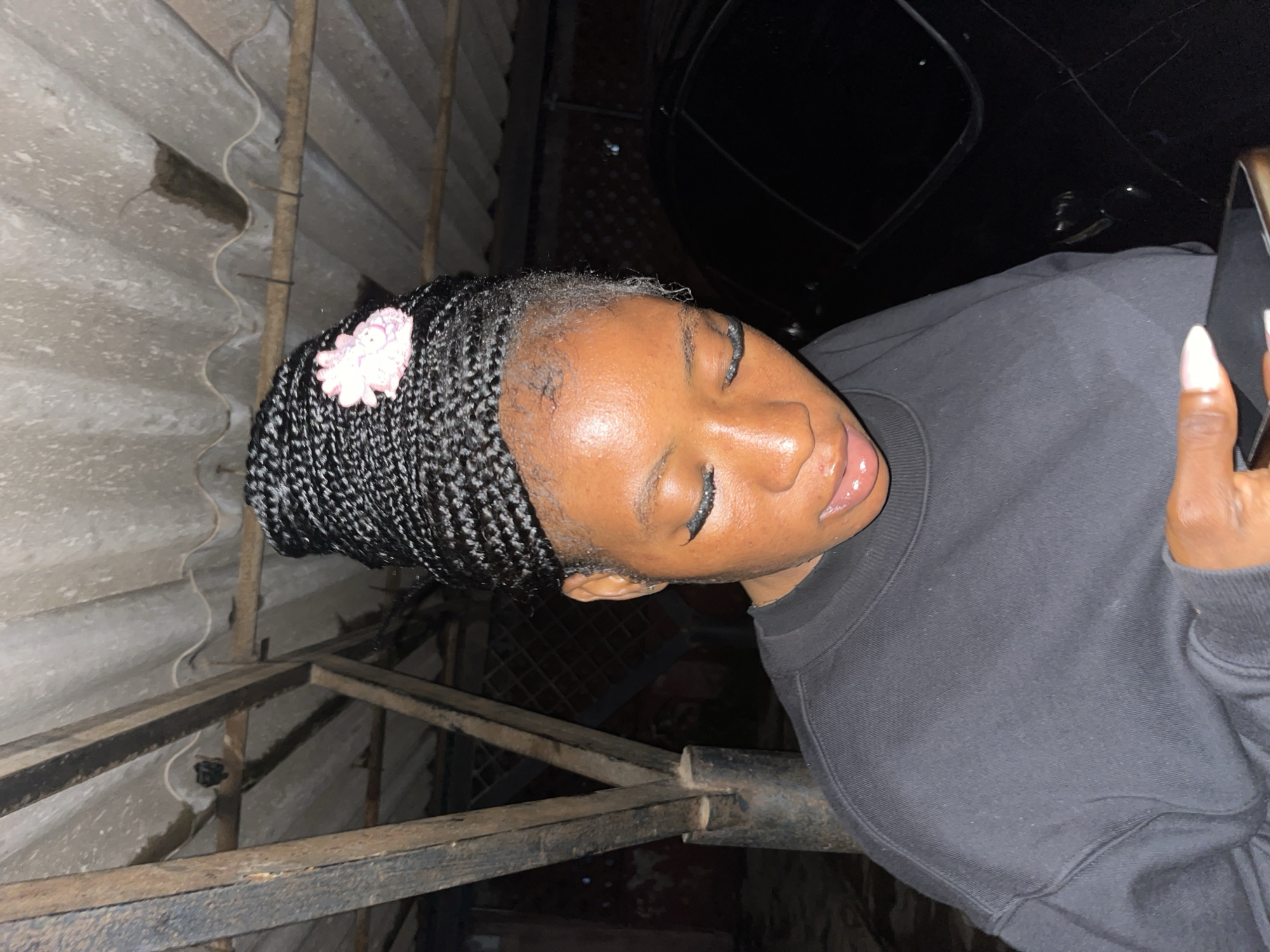A medium shot shows a young Black woman with braided hair styled in a high bun and adorned with a pink flower accessory, looking down at a smartphone she holds in her hands. She is wearing a black crewneck sweatshirt. The background is dimly lit, revealing a corrugated metal roof supported by wooden beams. The scene appears to be an outdoor, covered area, possibly a carport or workshop, in Jos, Nigeria. A dark-colored vehicle is partially visible to the right.