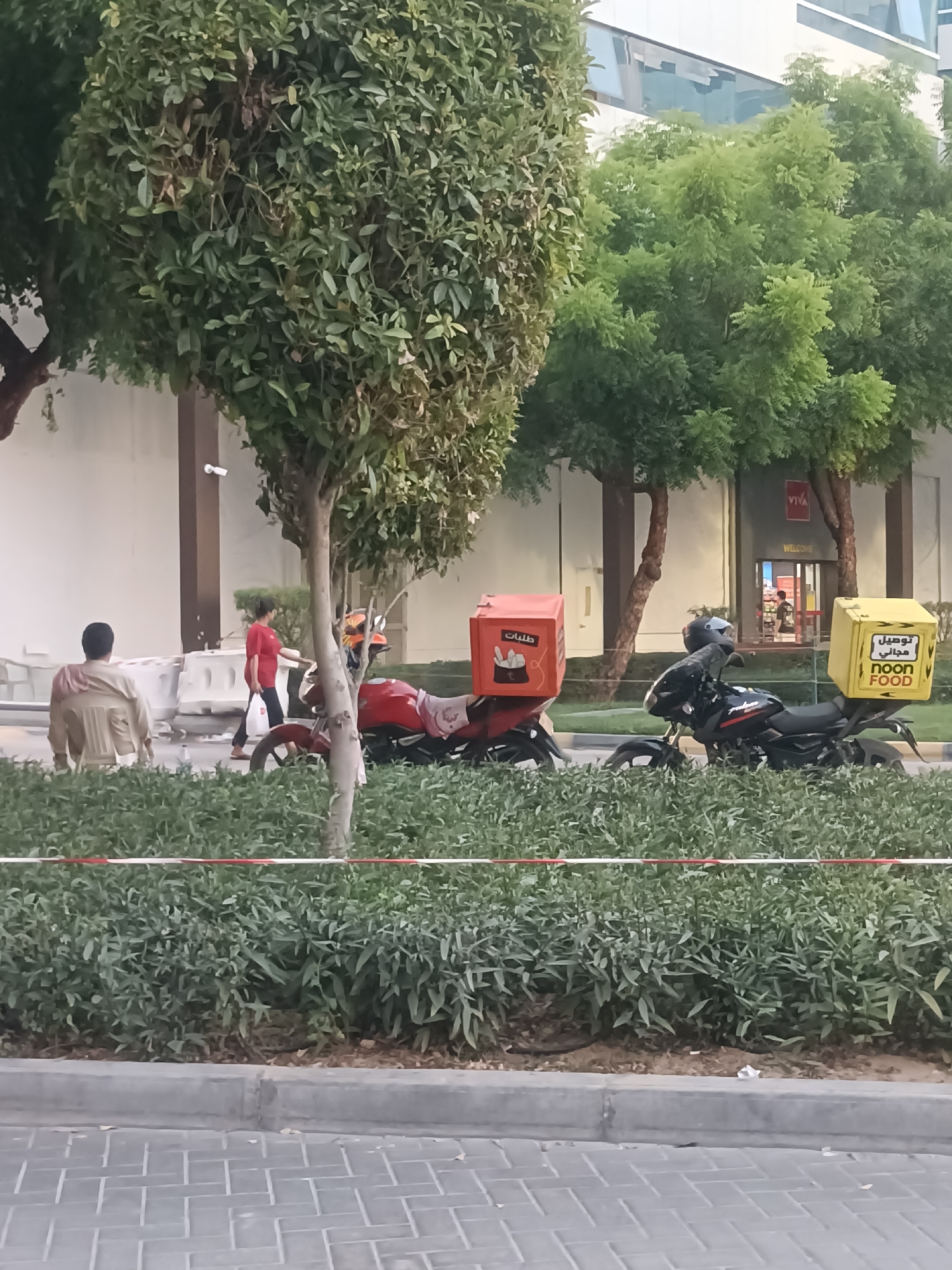 The image captures a street scene in Dubai, United Arab Emirates, likely near a commercial area, given the presence of a building with large glass windows and a storefront. In the foreground, two motorcycles are parked side-by-side, each equipped with large food delivery boxes. The motorcycle on the left has a red box with Arabic writing and a graphic of a food item. The motorcycle on the right has a yellow box with "NOON FOOD" written in English and Arabic. Both motorcycles are black.
Behind the motorcycles, a person in a red shirt and dark pants is walking away from the camera, carrying a bag. To the left of this person, a person in a light-colored, possibly beige, outfit is seated, facing away from the camera.
The environment features lush green trees and bushes, creating a somewhat park-like setting along the street. A paved sidewalk or walkway is visible in the foreground, with a curb and a patterned brick surface. In the background, a modern building with reflective glass windows is visible. A security camera is mounted on a pillar near the seated individual. The lighting suggests it is daytime, possibly late afternoon or early morning, with soft shadows.