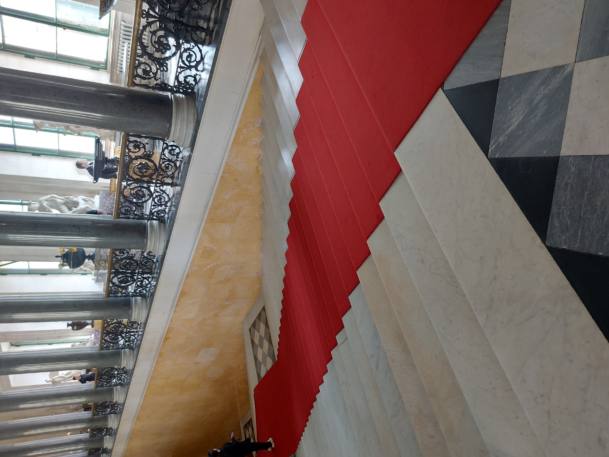 The image shows a view of a staircase in a grand building. A red carpet runs up the center of the stairs, which are made of white marble. The floor at the top of the stairs is checkered in black and white. To the left of the stairs is a wall with a yellow marble pattern and a series of gray columns with ornate black railings. People can be seen in the background, and statues are visible through the windows behind the columns. The style of architecture suggests this is located in Saint Petersburg, Russia.