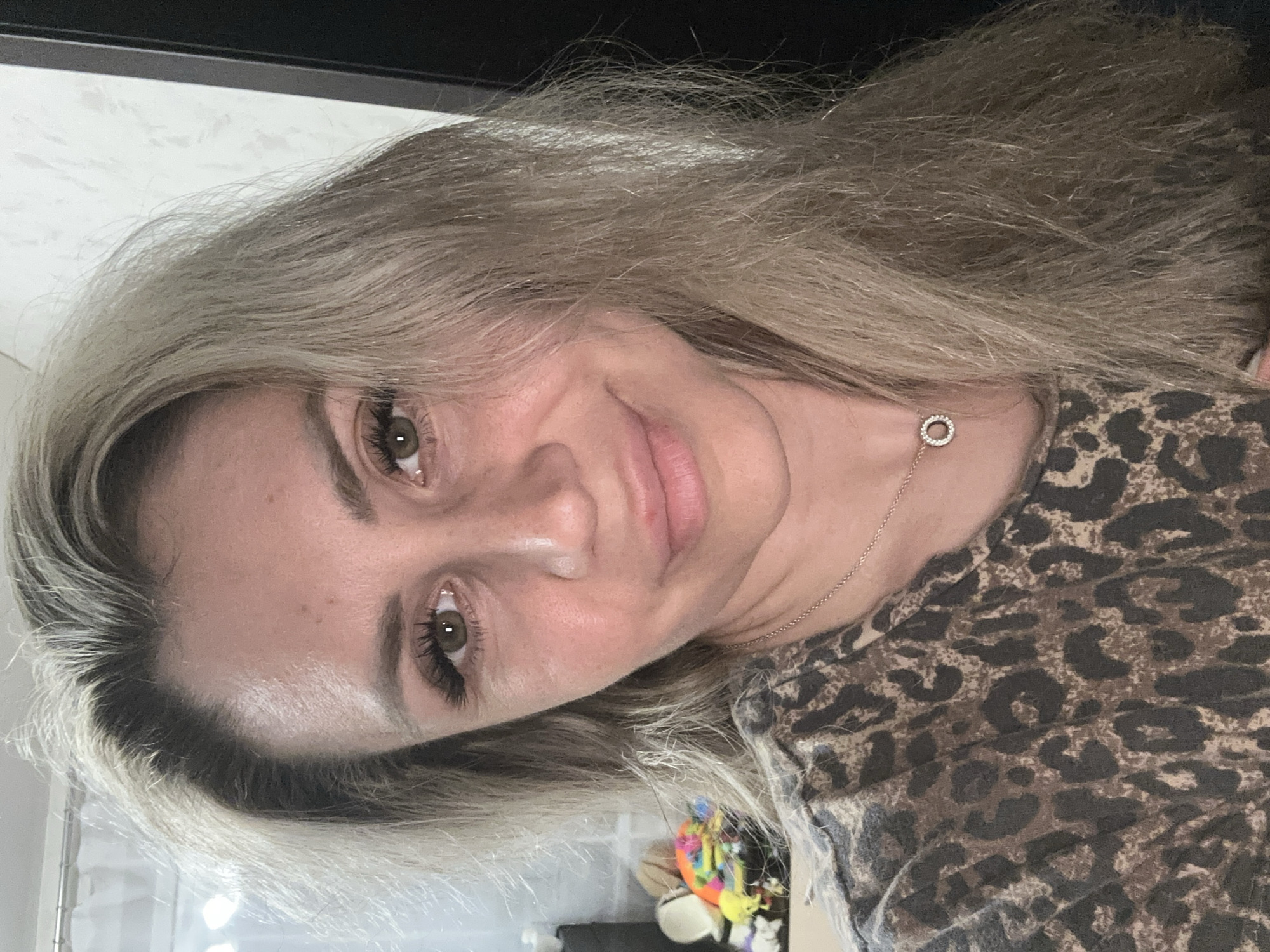 A close-up, head-and-shoulders selfie shows a woman with fair skin, blonde hair, and green eyes. She is wearing a leopard-print top and a delicate necklace with a circular pendant. The background is indistinct, but appears to be an indoor setting with a window and a dark surface to the right. The lighting suggests it is daytime. The image is taken at a slightly upward angle, focusing on the woman's face. There is no discernible location in Novoaltaysk, Russia.
