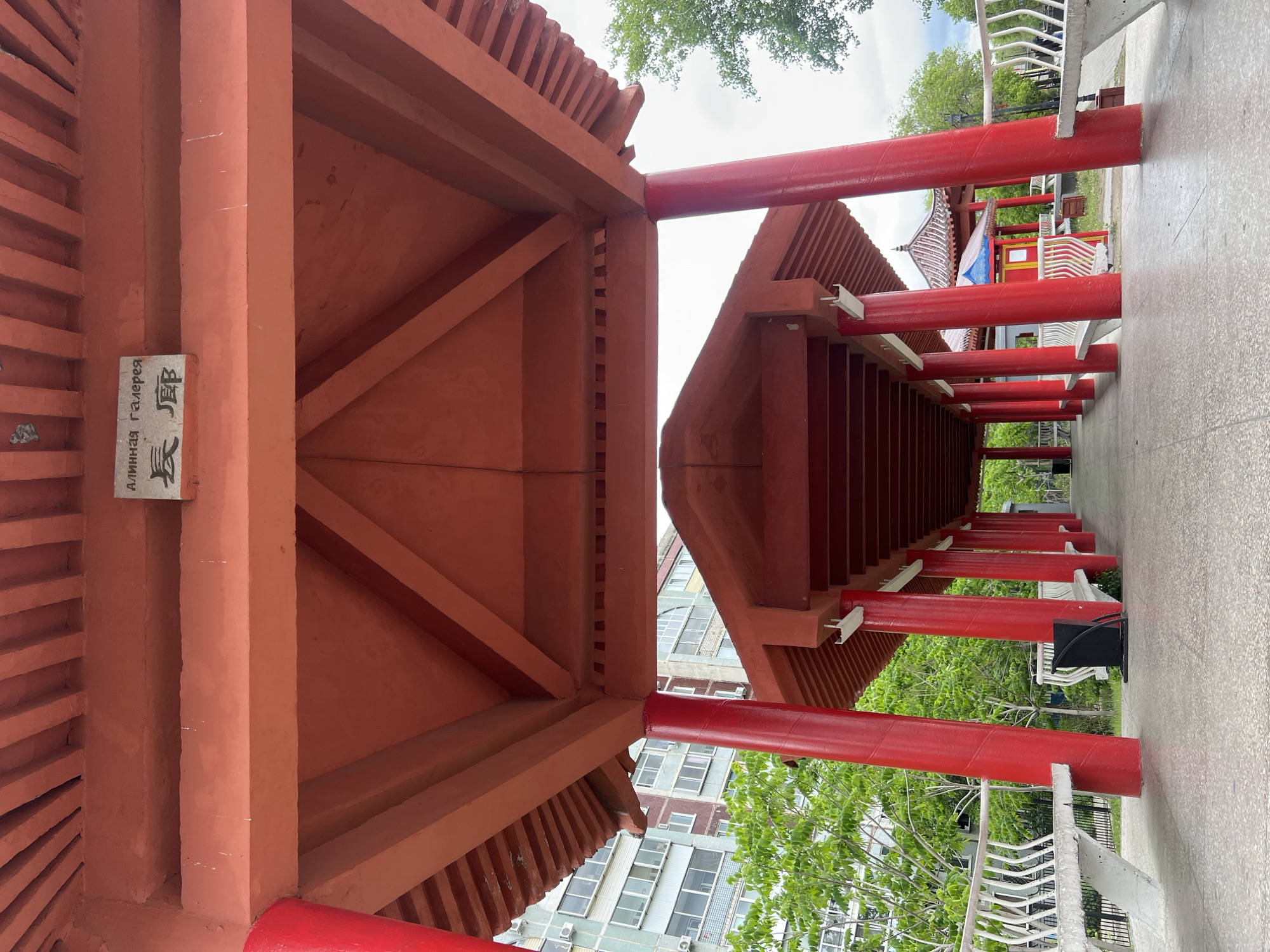 The image shows a red, pagoda-style structure, possibly a walkway, likely located in a public park or garden, as suggested by the surrounding greenery and architecture of other buildings in the background. The perspective is from within the structure, looking lengthwise down the passage. Red columns support the pagoda roof. The architecture is possibly inspired by Chinese or East Asian styles. A sign with writing in a non-Latin alphabet is visible on one of the vertical supports.