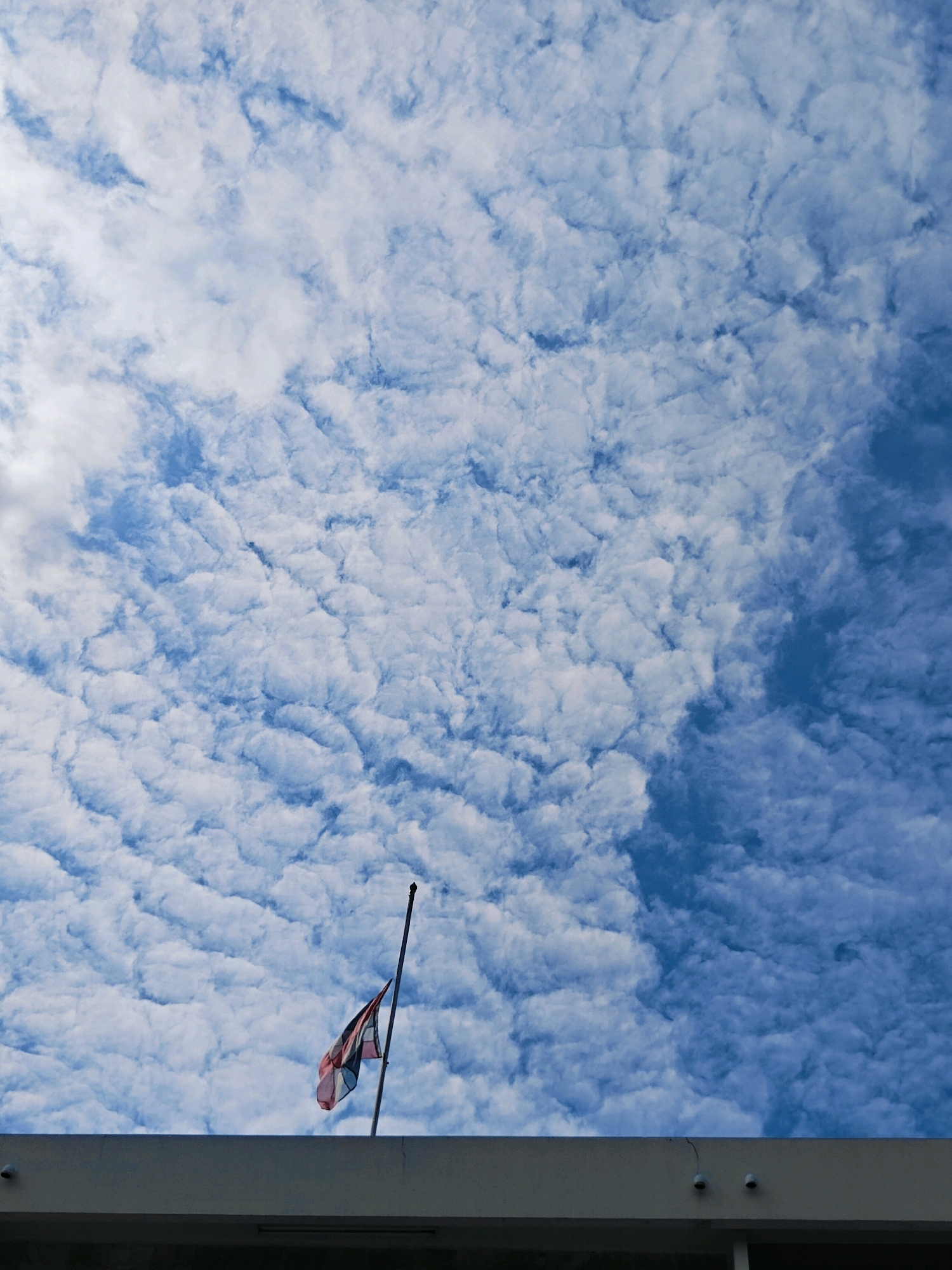 A low-angle outdoor shot shows a section of a building's concrete facade in the foreground. Above the facade, a blue sky is filled with a dense, textured layer of white, puffy clouds. A flag pole extends upwards from the facade, with a flag flying at half-mast. The flag is partially visible and appears to be the flag of Thailand, with red, white, and blue stripes. The scene is likely captured during daylight hours.