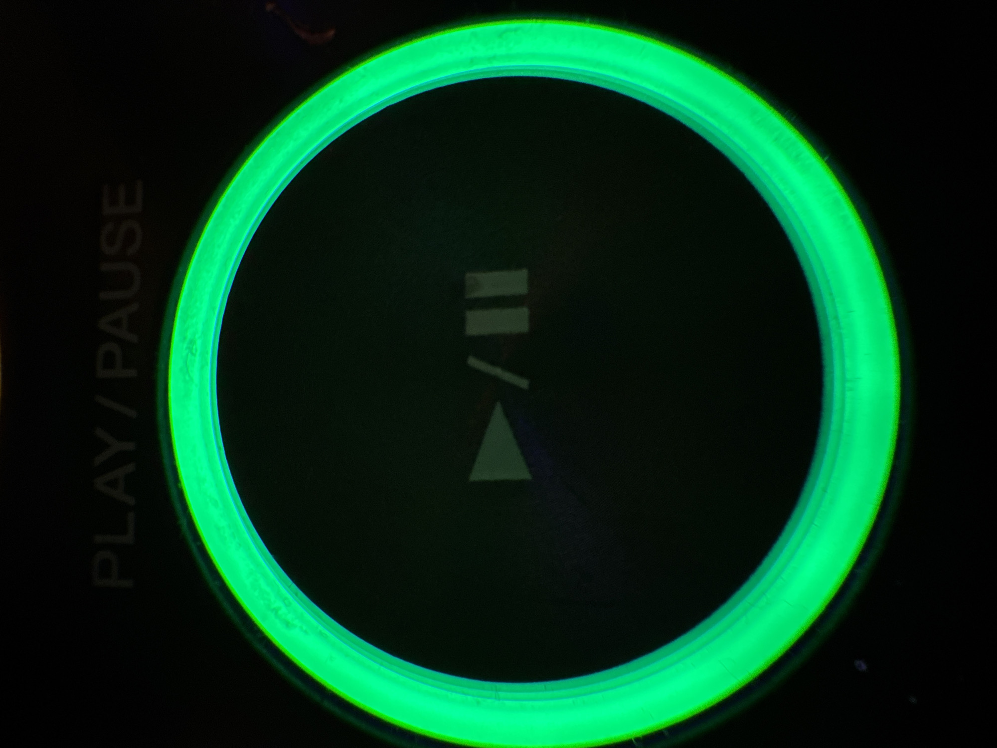 A close-up shot focuses on a circular, green backlit button labeled "PLAY/PAUSE" situated on a dark background. The button's center displays a play/pause symbol. The location is unidentifiable from the visual information provided.