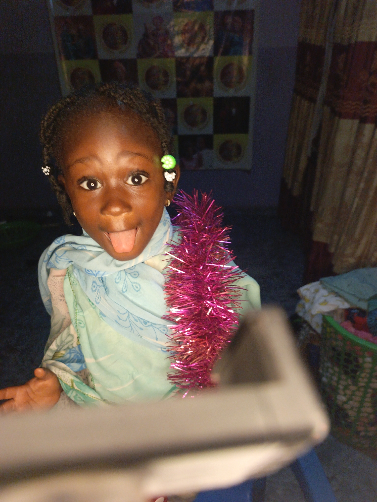 A young girl with dark skin and braided hair sticks her tongue out at the camera. She is wearing a light blue and green patterned scarf and a pink tinsel garland is draped around her neck. She has small white and green beads in her hair. In the background, a checkered tapestry hangs on the wall, and a beige curtain is visible on the right. A green plastic basket filled with items is also in the background. The scene appears to be indoors, possibly in a home in Akwanga, Nigeria.