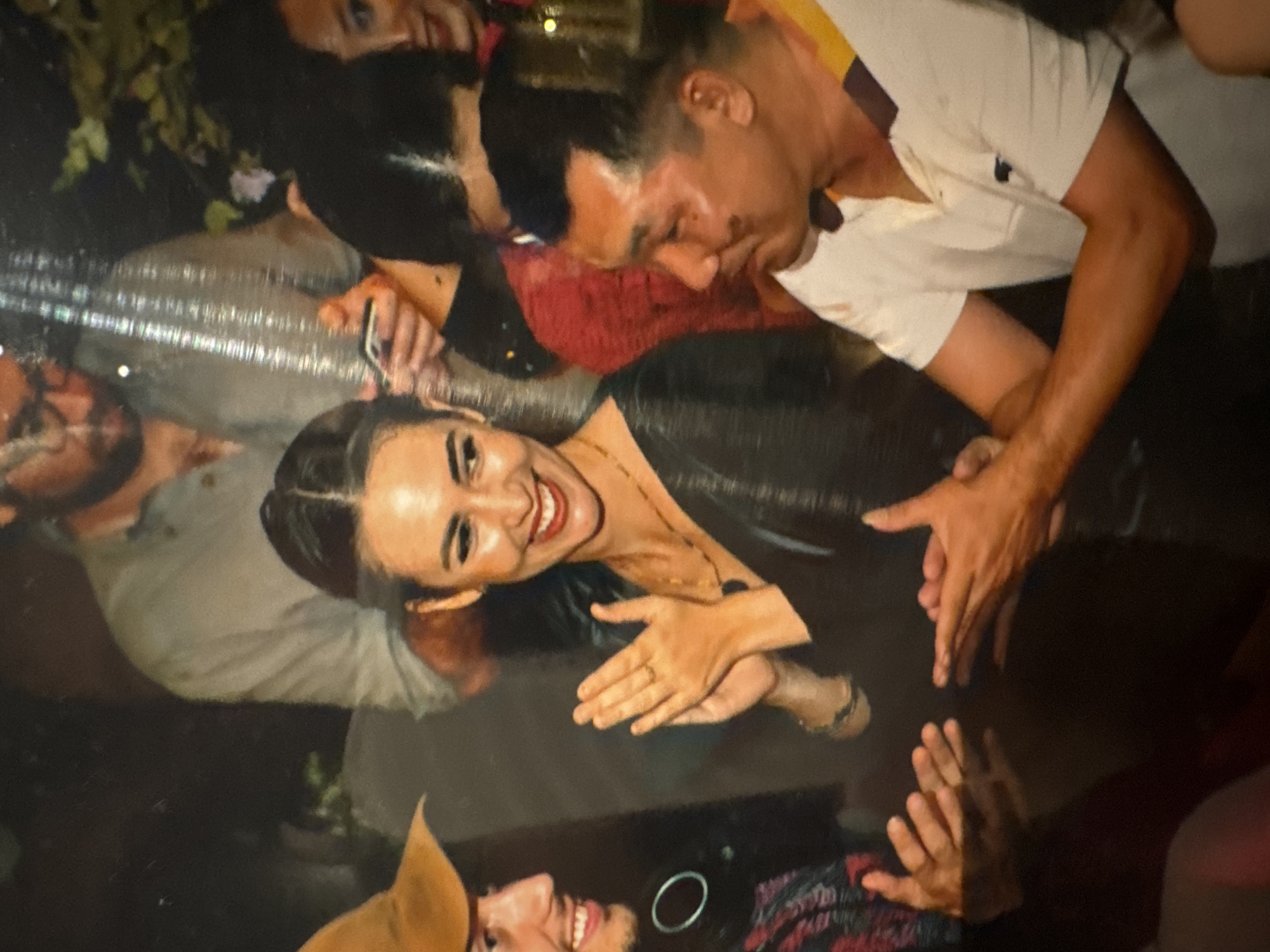 The image captures a group of people in what appears to be an indoor social gathering. A woman in the center is smiling broadly and clapping her hands, wearing a dark, shimmering dress and a necklace. To her left, a man with a tan hat and headphones is also smiling and clapping. To the woman's right, a man in a white polo shirt with a dark collar is looking to the side, his hands extended. In the background, other individuals are visible, some partially obscured. The lighting is dim, suggesting an evening event. The location is not identifiable as Hà Nội, Vietnam based on visual cues in the image.
