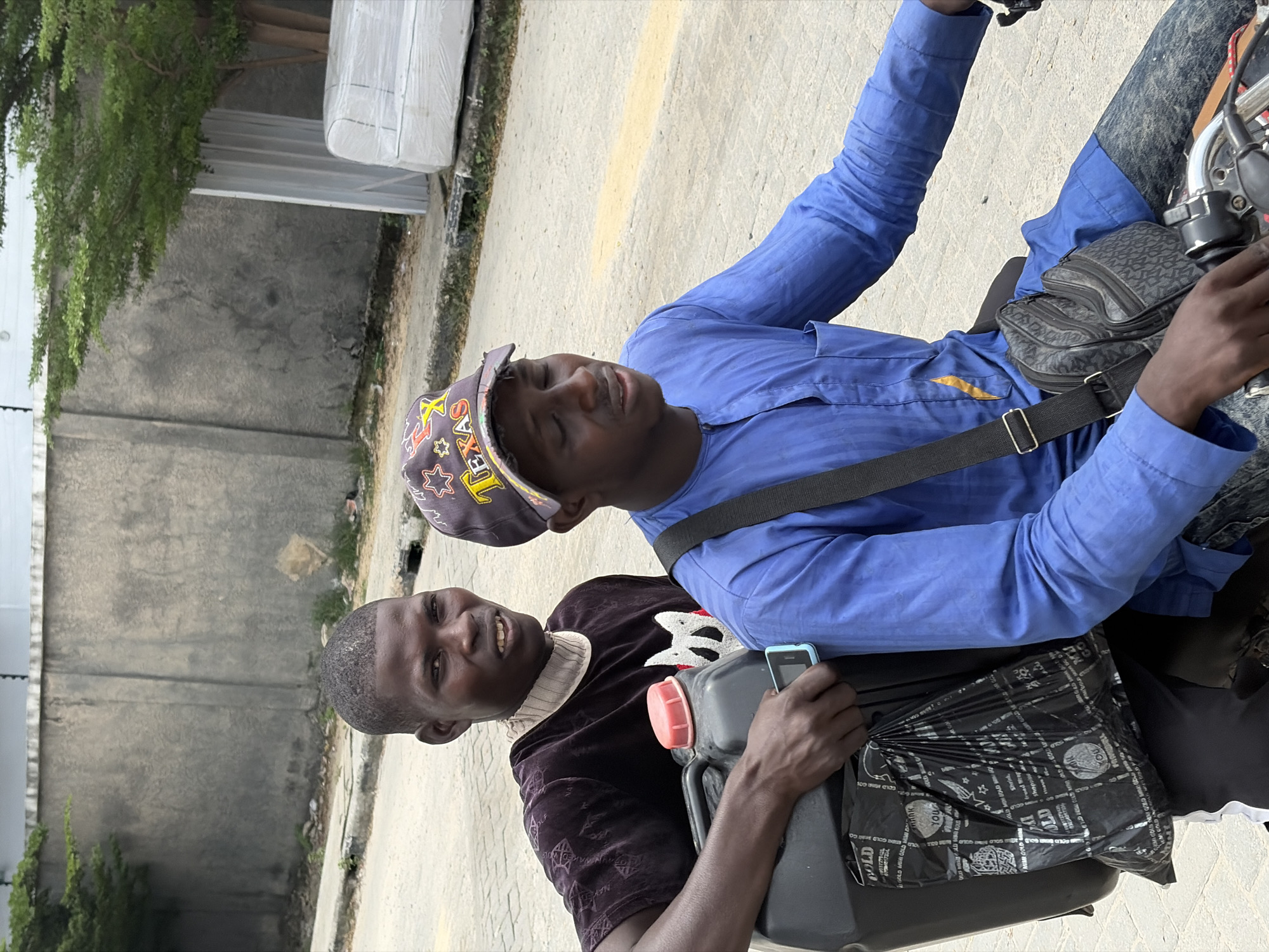 Two men on a motorcycle in Igboefon, Nigeria. One is wearing a blue shirt and a "TEXAS" baseball cap, while the other is wearing a dark patterned shirt and holding a black plastic bag and a black container with a red cap. They are on a paved street next to a building with a concrete wall and some vegetation. The man in the back is holding a small cellphone.