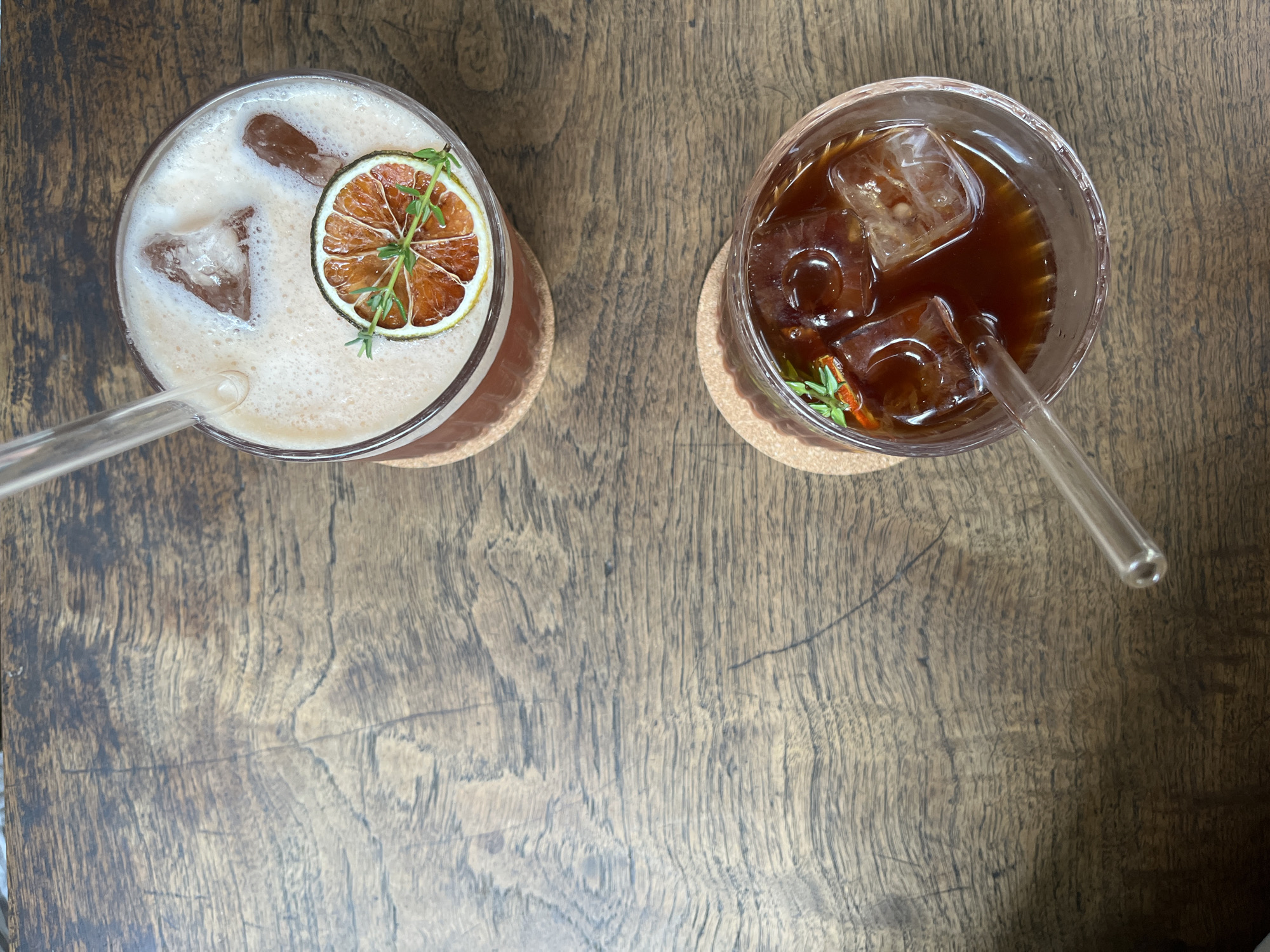 Two glasses of cold beverages are placed on a wooden surface, likely a table. The top glass contains a frothy, light-colored drink with ice cubes and is garnished with a slice of dried orange and a sprig of thyme. A clear straw extends from the drink. Below it, the second glass holds a dark-colored beverage, also filled with ice cubes and garnished with thyme and what appears to be a small red chili. A clear straw is present in this glass as well. The wooden surface exhibits a prominent grain pattern. The scene suggests a cafe or restaurant setting in Thủ Đức, Vietnam.