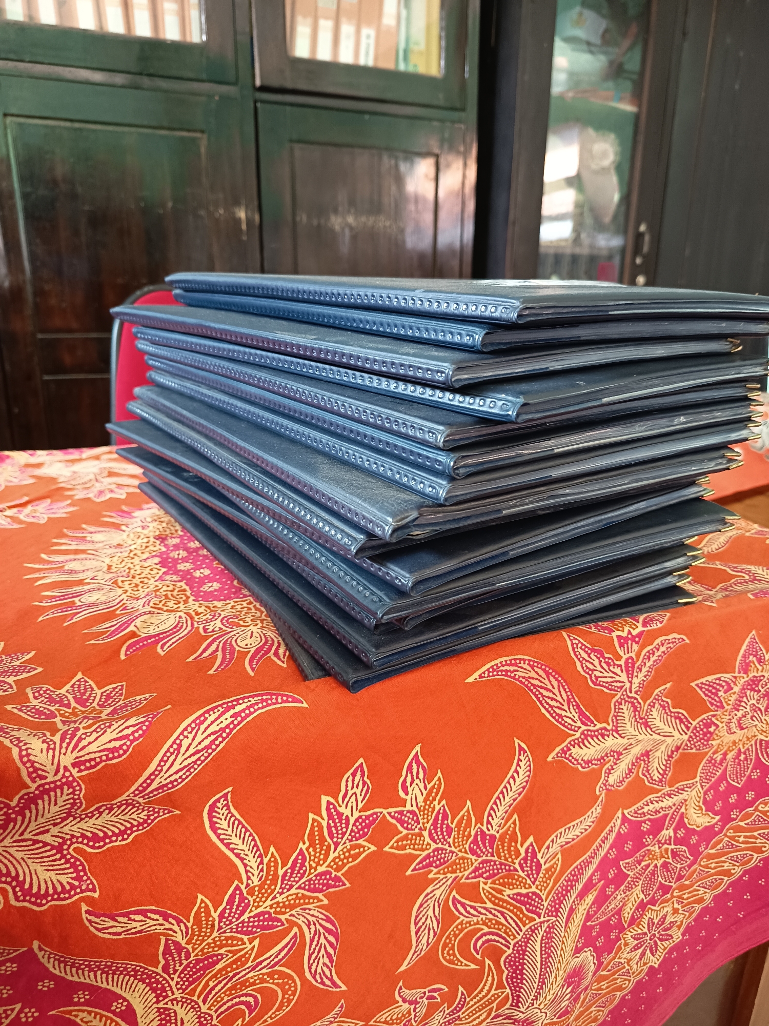 A stack of blue-covered folders sits on a table covered with an orange and pink floral batik cloth. In the background, a dark wooden cabinet with glass doors is visible, and inside the cabinet, shelves filled with books can be seen. The scene appears to be indoors, possibly in an office or library in Widoro, Indonesia.