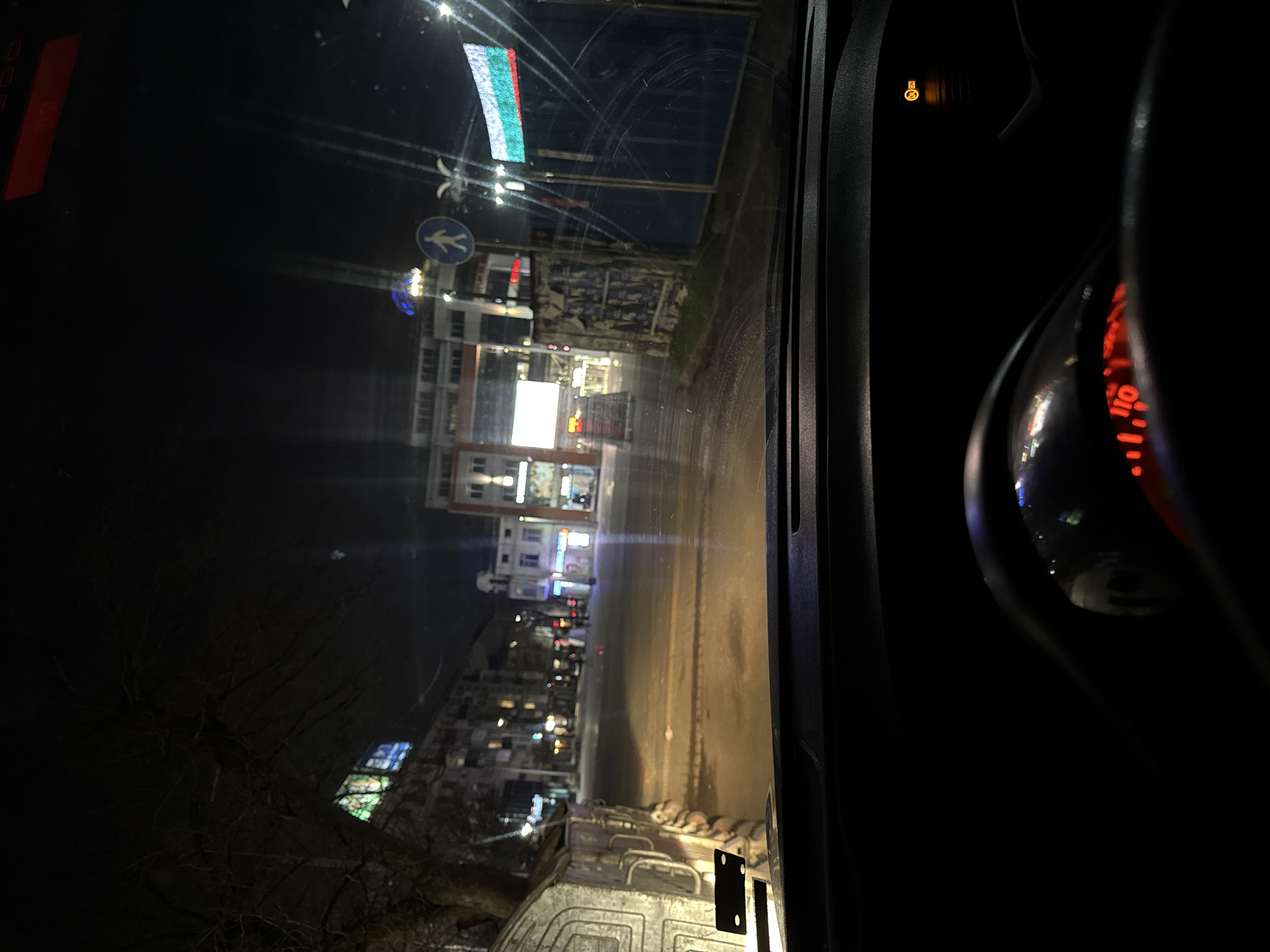 The image shows a view through a car windshield at night. The car is on a street in Burgas, Bulgaria. The street is lined with buildings and lit by streetlights. There is a Bulgarian flag visible. The car's dashboard and speedometer are partially visible. A truck is immediately outside the car.