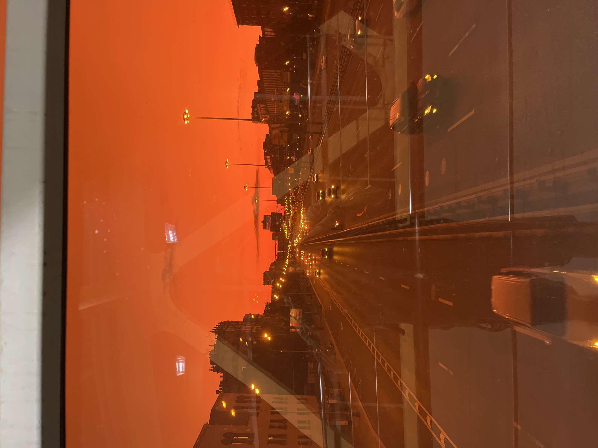 The image captures a view through a window onto a highway at dusk. The sky is an intense orange, suggesting a phenomenon such as a dust storm or unusual atmospheric conditions. Buildings line the highway, illuminated by streetlights and building lights. Cars are visible on the highway, their headlights and taillights glowing. The reflection on the window is prominent, obscuring parts of the scene and showing a distorted view of the interior or another scene. The overall impression is one of a busy urban environment under an exceptionally colored sky. This scene is located in Khimki, Russia, specifically on the Leningradskoye Highway, known for its wide lanes and surrounding urban development.