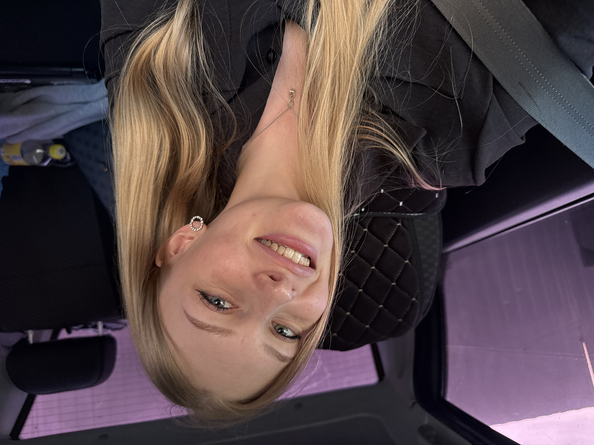 A young woman with long blonde hair and blue eyes is smiling at the camera. She is wearing a black blazer and a seatbelt. She has a silver earring and necklace. She appears to be inside a car. Based on the location information provided, the scene is likely in or near the city of Кондратово, Russia. The details do not provide any specific context to indicate specific actions or interactions taking place, only the depiction of the subject within the vehicle.