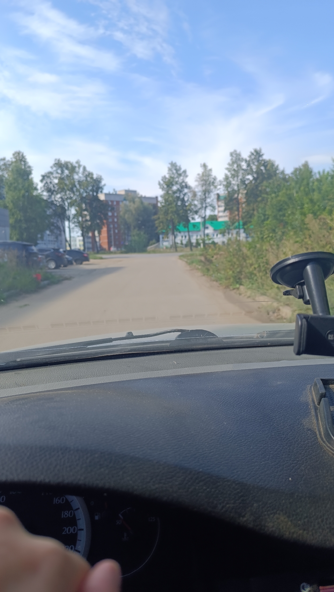 The image, taken from inside a vehicle in Chaikovsky, Russia, shows a view through the windshield of a paved road leading into a residential area. On the left side of the road, several parked cars are visible adjacent to a grassy area. Further ahead, multi-story apartment buildings with red brick and white facades, interspersed with trees, line the street. Green trees and foliage are abundant on both sides of the road. The sky above is blue with scattered white clouds. In the foreground, the dashboard of the car is visible, including a speedometer with markings up to 200, a fuel gauge, and a hand on the steering wheel. A suction cup mount for a device is attached to the windshield on the right side. The scene suggests a daytime setting with clear weather.
