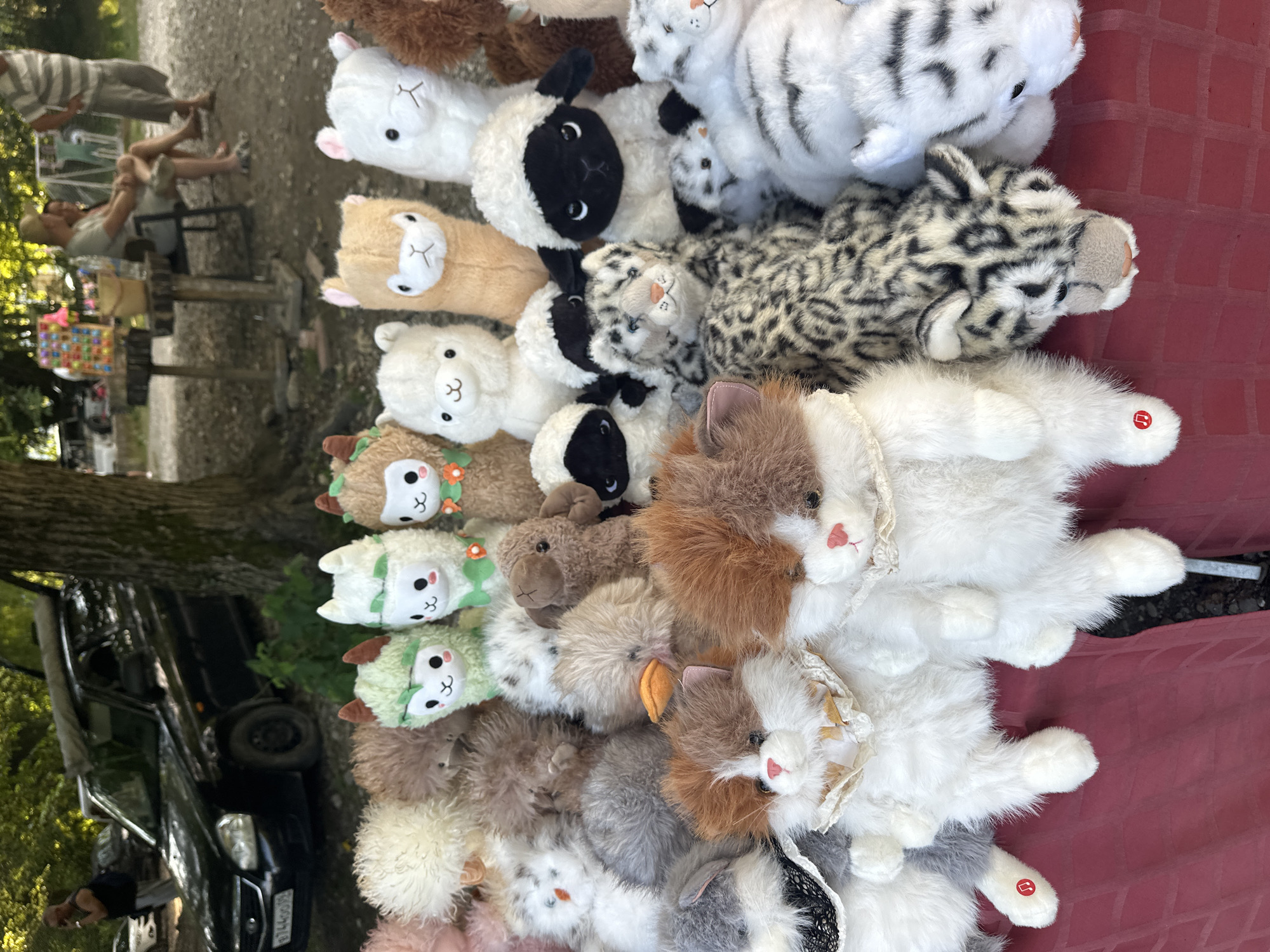 A collection of stuffed animals, including alpacas, sheep, cats, and a leopard, are displayed on a red tablecloth. The stuffed animals are in various sizes and colors. In the background, a black car is parked, and several people are visible. The setting appears to be an outdoor market or roadside stall in Bol'shoi Kichmai, Russia. The stuffed animals are the primary focus of the image, with the background providing context for the location.